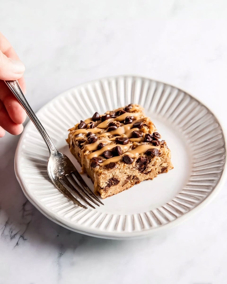 A square bar with a light tan base filled with small dark brown chocolate chips on top and inside, drizzled with a light brown sauce in uneven lines. It sits on a round white plate with raised lines along the edge. A silver fork is placed on the plate, with a woman's hand holding the fork on the left side. The background is a white marbled surface. photo taken with an iphone --ar 4:5 --v 7