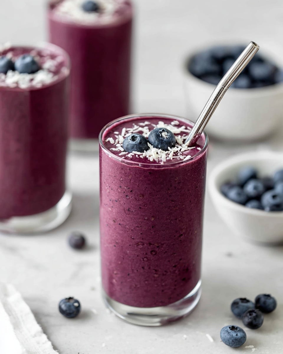 The image shows a tall clear glass filled with a thick, deep purple smoothie. On top of the smoothie are two whole blueberries and a sprinkling of white coconut flakes. A silver metal straw is placed inside the drink, angled slightly towards the right. In the background, there are two more glasses filled with the same smoothie, one fully visible and one partially visible, both topped with blueberries and coconut flakes. Scattered fresh blueberries are placed on a white marbled surface, and a white bowl filled with blueberries is slightly out of focus in the upper right corner. The photo has soft lighting and a clean, fresh look. photo taken with an iphone --ar 4:5 --v 7