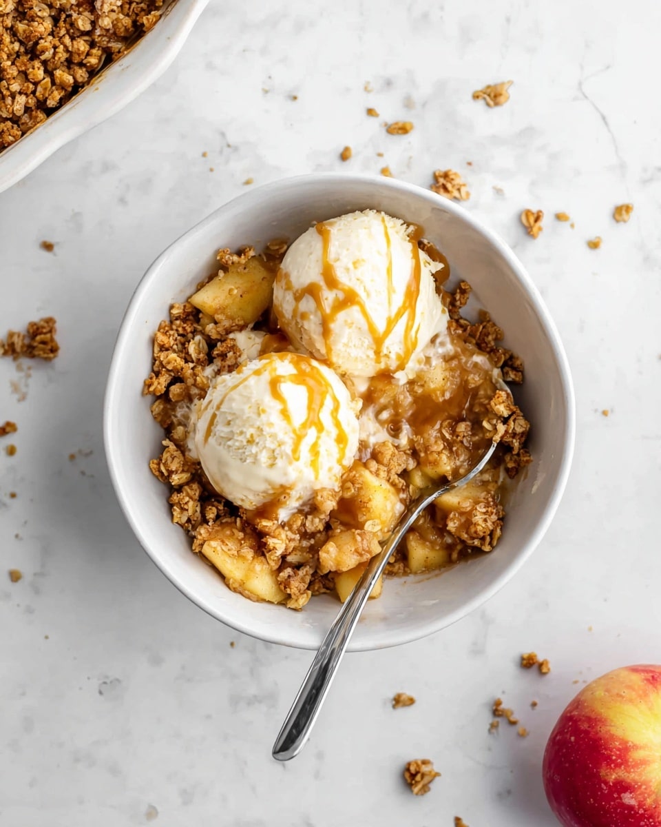 A white bowl filled with a layered dessert, the bottom layer contains golden brown oat crumble with visible chunks of cooked apple pieces. On top, there are two scoops of creamy white vanilla ice cream drizzled lightly with caramel sauce. A silver spoon rests inside the bowl, partially submerged in the dessert. The bowl is placed on a white marbled surface with crumbs scattered around and a halved red apple to the right. Photo taken with an iphone --ar 4:5 --v 7