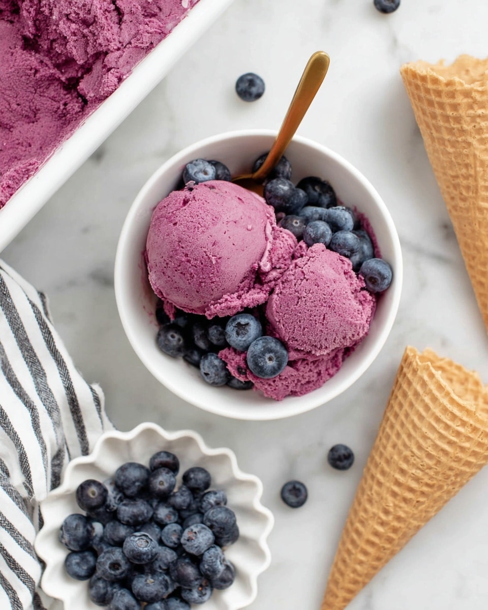 A white bowl filled with two scoops of bright purple frozen dessert, showing a creamy and slightly rough texture, is topped with several fresh, plump blueberries scattered on and around the dessert. Next to the bowl, there is a gold spoon resting inside the dessert. To the right of the bowl, two empty waffle cones lay on a white marbled surface. Below the bowl, a small white scalloped dish holds a pile of fresh blueberries. A white cloth with black stripes is partially visible on the left side of the image. Part of a larger container with the same purple dessert is seen at the top left corner. photo taken with an iphone --ar 4:5 --v 7