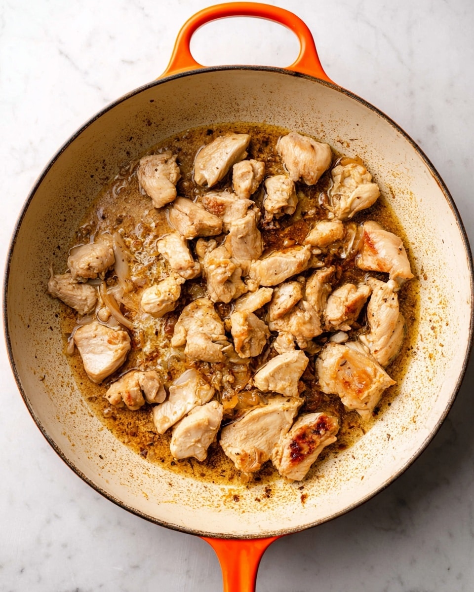 A large, round white pan with an orange handle shows several pieces of browned chicken cooking in the center. The chicken pieces vary in size and have a light golden color with some parts appearing slightly crispy. The pan surface has a mix of oil and browned bits, giving a textured, slightly oily look. The pan sits on a white marbled surface. photo taken with an iphone --ar 4:5 --v 7