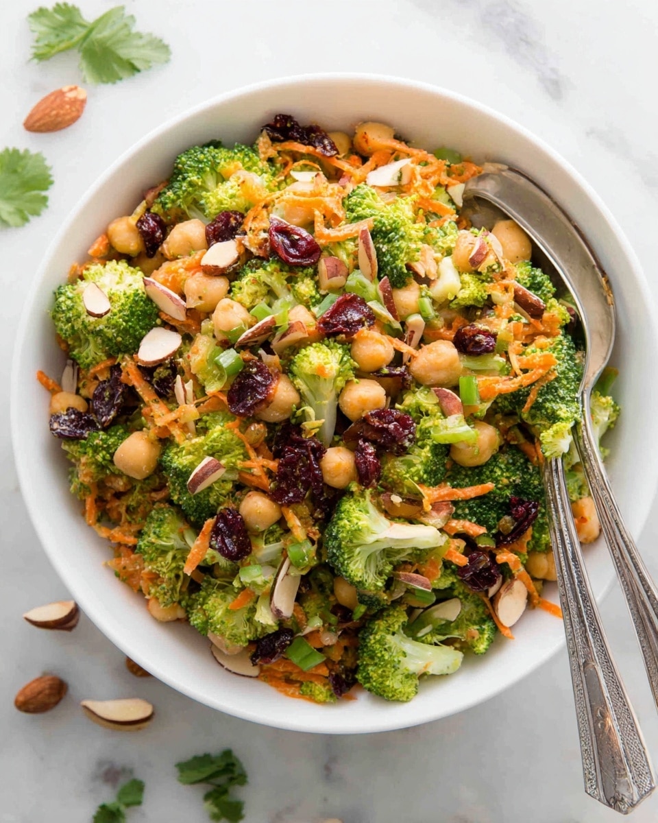 A white bowl filled with a colorful salad that has three main layers: bright green broccoli florets cut into small pieces forming the base, a middle layer of thin orange shredded carrots mixed with light beige chickpeas, and a top layer sprinkled with dark red dried cranberries and light brown almond slices. The salad is mixed with small green onion pieces and appears lightly coated with a creamy dressing, giving a slight shine to the vegetables. Two metal spoons rest inside the bowl, and there are a few almond slices and green cilantro leaves scattered around the bowl on a white marbled surface. Photo taken with an iphone --ar 4:5 --v 7