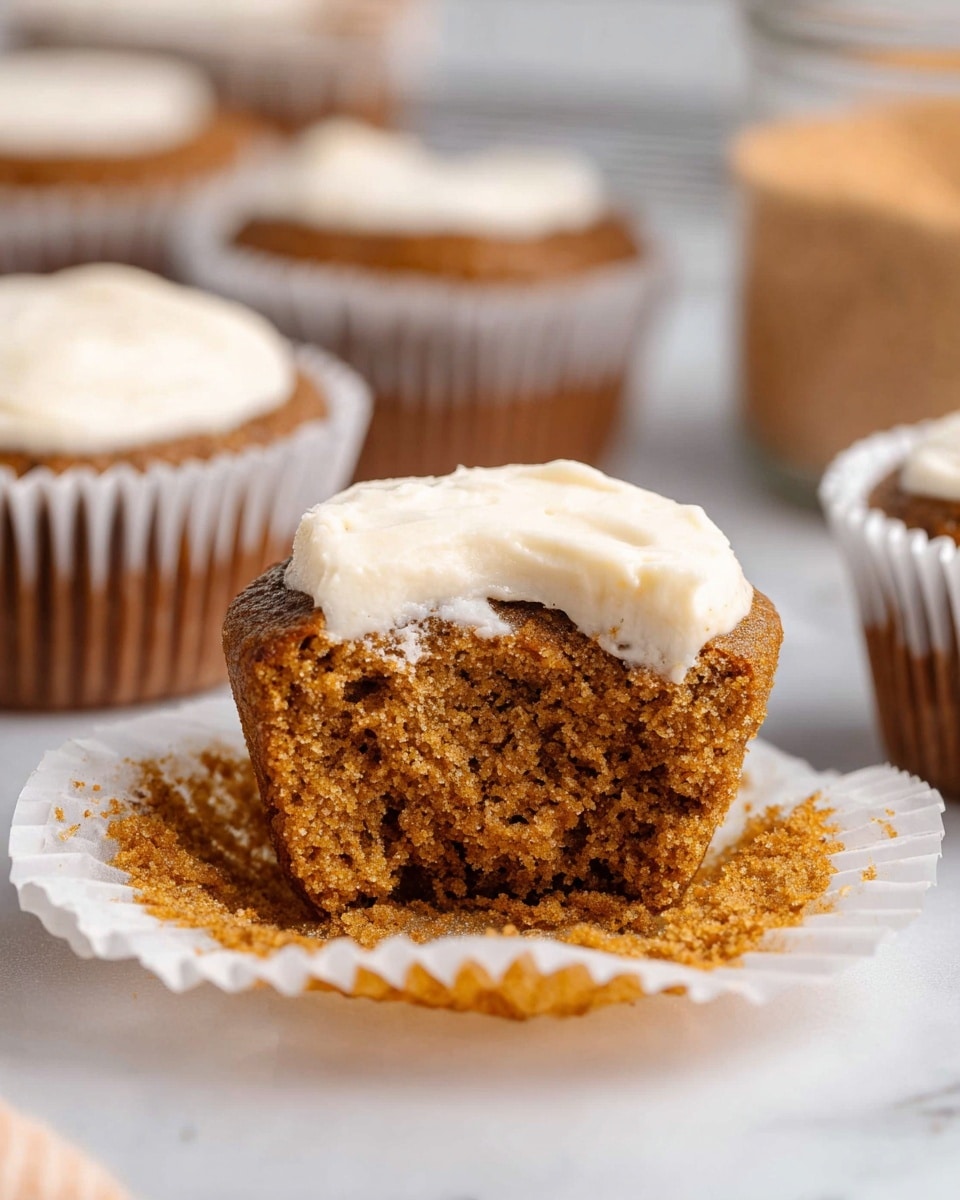 A close-up of a single small muffin with a light brown, moist, and soft texture, partially eaten from the front to show the crumb inside. The muffin has a white, creamy frosting layer on top, centered and thick, contrasting with the brown cake. It sits in a white paper muffin liner with ridges, placed on a white marbled surface. In the blurred background, there are other muffins in similar white paper liners and a glass jar containing light brown powder, all softly out of focus. photo taken with an iphone --ar 4:5 --v 7