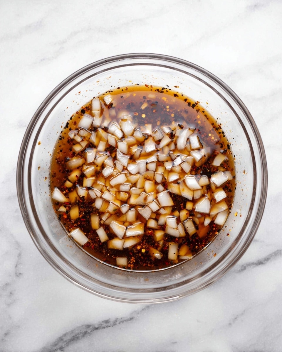 A clear glass bowl on a white marbled surface holds a liquid mixture. The liquid is dark brown with a slightly oily texture and contains many small, diced white onion pieces floating throughout. The onions are spread evenly across the surface, creating a contrast with the brown liquid, and tiny black and reddish spice particles are visible in the mixture. The bowl is filled about halfway, showing the layers clearly through the transparent glass. photo taken with an iphone --ar 4:5 --v 7