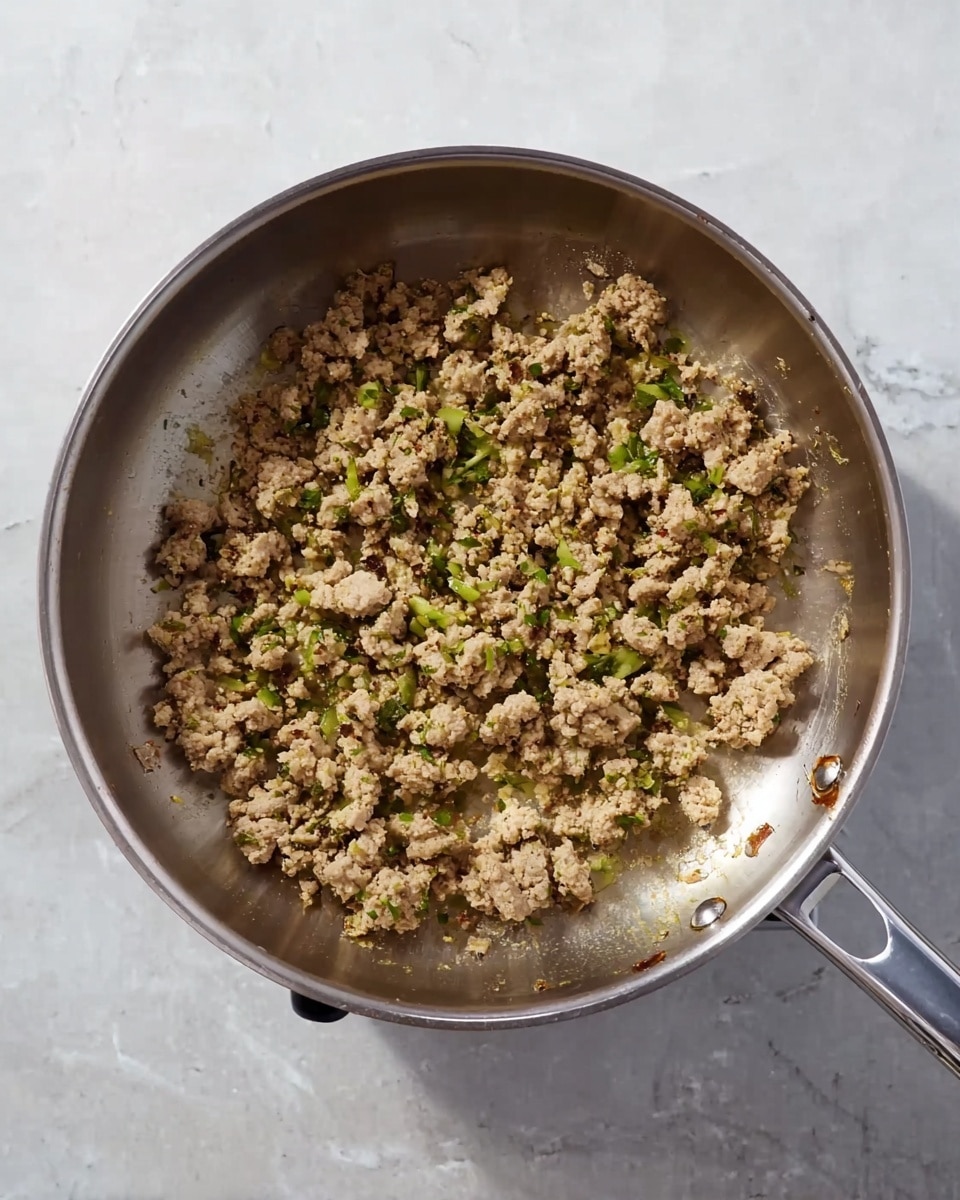 A metal pan is seen from above on a shiny silver stove, filled with cooked ground meat mixed with small pieces of green herbs or vegetables spread unevenly inside. The ground meat has a light brown and beige color with a crumbly texture, showing some browned bits near the edges. The background is a white marbled surface with subtle grey veins. photo taken with an iphone --ar 4:5 --v 7