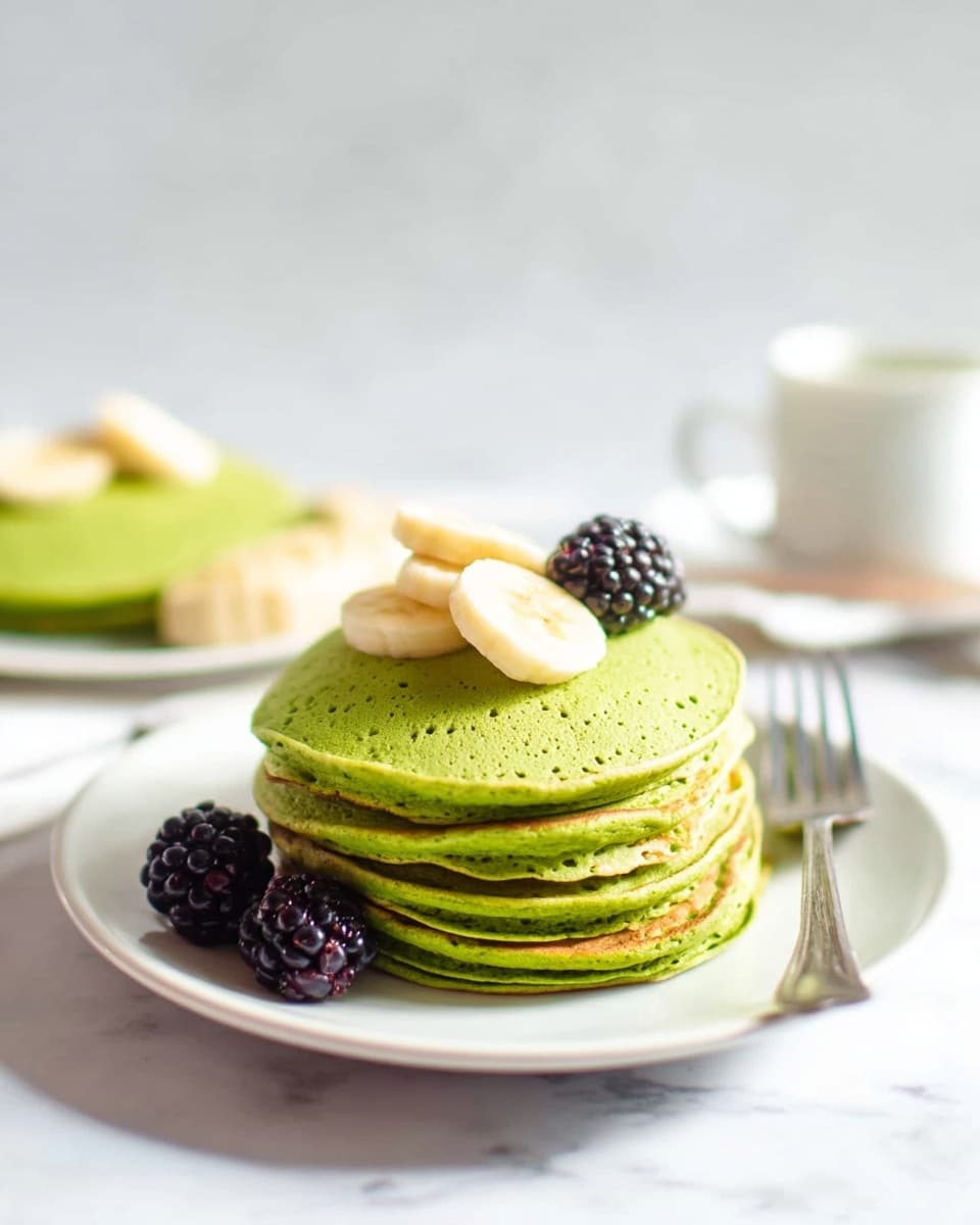 A stack of five green pancakes sits in the center of a white plate on a white marbled surface. The pancakes have a fluffy texture with tiny holes visible on their edges. On top of the stack, there are three slices of banana neatly placed. Two blackberries rest on the plate beside the pancakes. A silver fork lies on the plate to the right of the pancakes. In the background, out of focus, there is another white plate with green pancakes and banana slices. Photo taken with an iphone --ar 4:5 --v 7