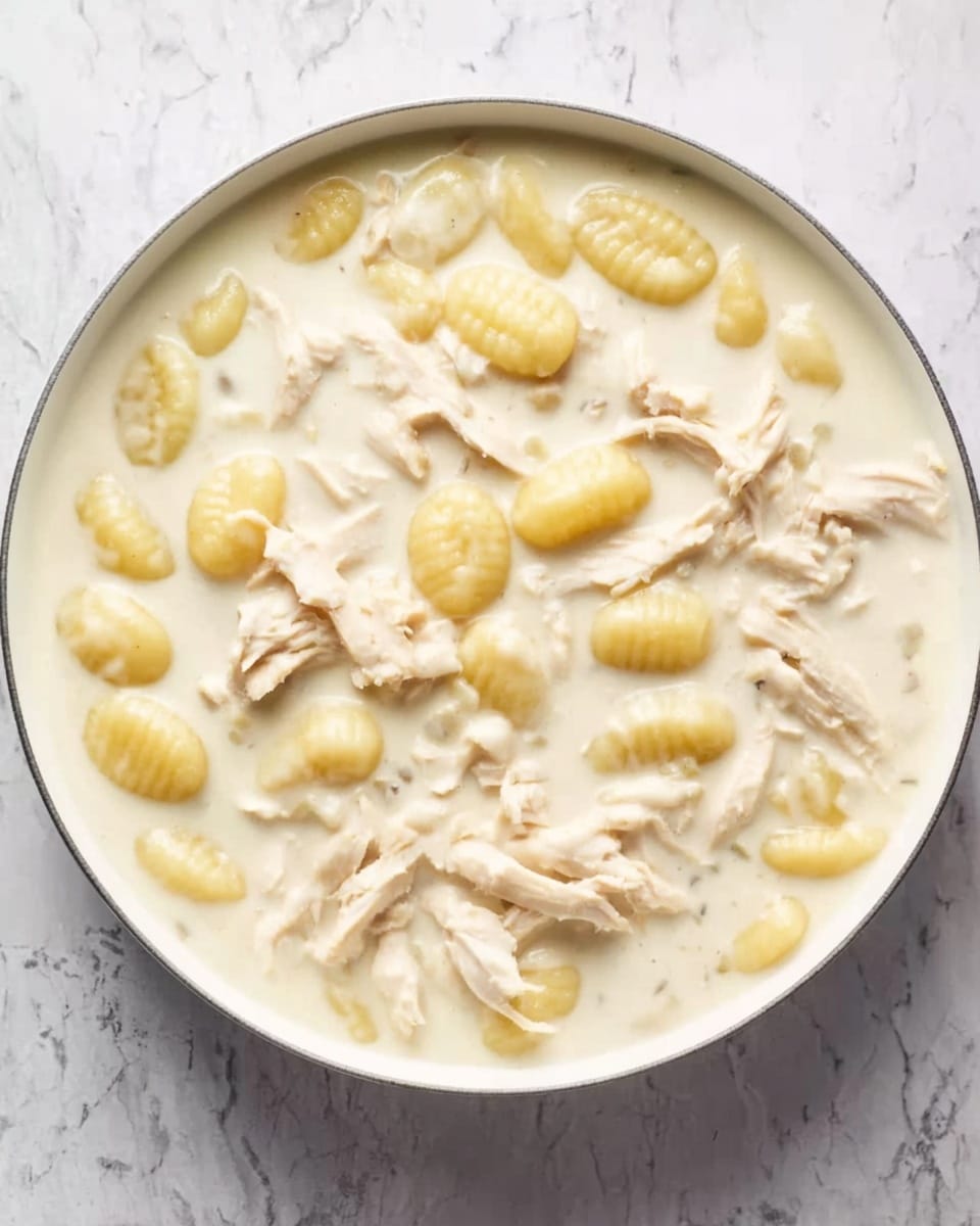 A white pan filled with a creamy white soup that covers most of the surface. Inside the soup, there are many light yellow gnocchi pieces spread evenly. Shredded pieces of white chicken are scattered throughout, mixed with the gnocchi and cream. The background is a white marbled texture. photo taken with an iphone --ar 4:5 --v 7