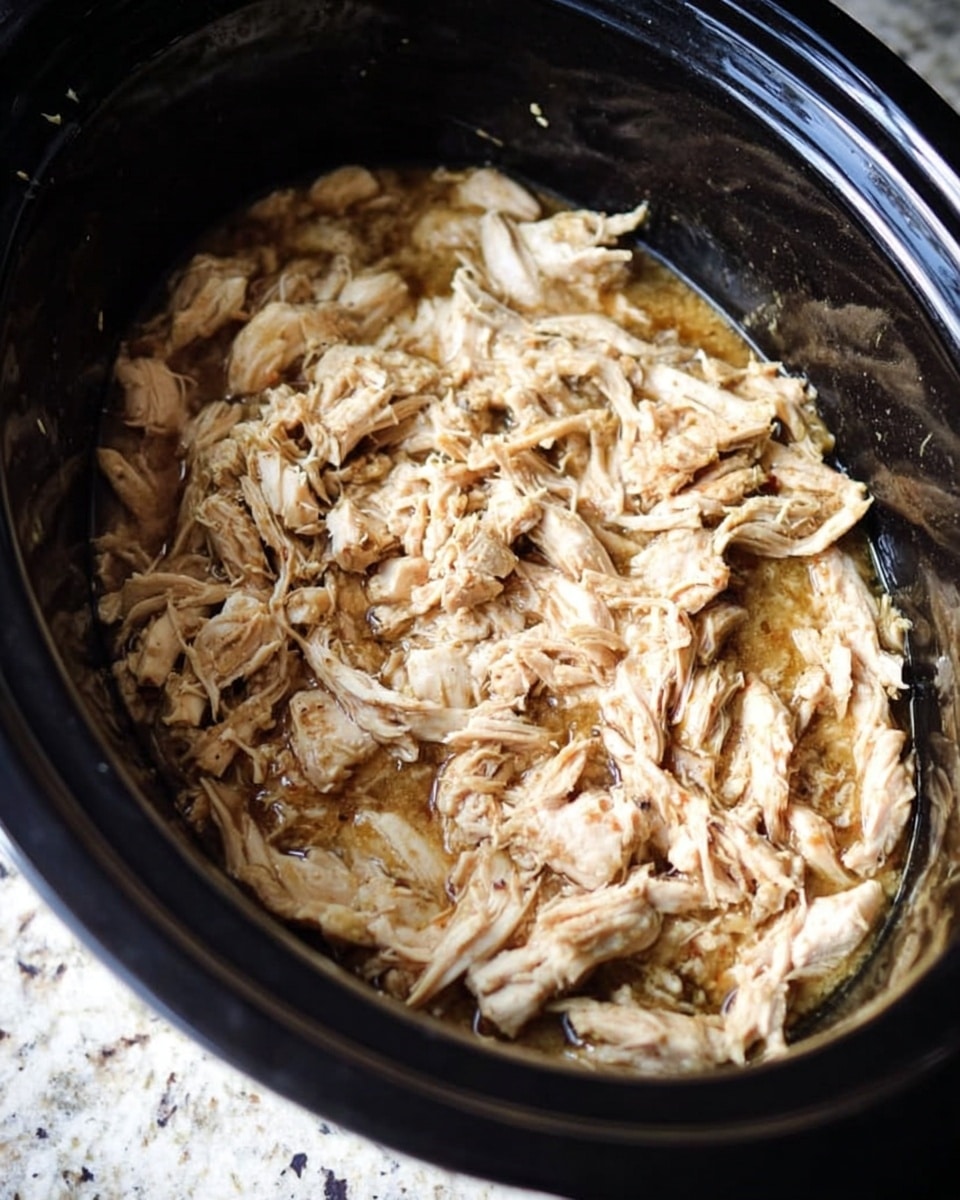 The image shows shredded white chicken cooked in a slow cooker with a light brown sauce covering it. The texture of the chicken looks soft and tender, with some pieces slightly darker from cooking. The sauce appears thin and slightly oily, coating the chicken evenly across one thick layer inside the black slow cooker. The surface beneath the cooker is white marbled texture. photo taken with an iphone --ar 4:5 --v 7