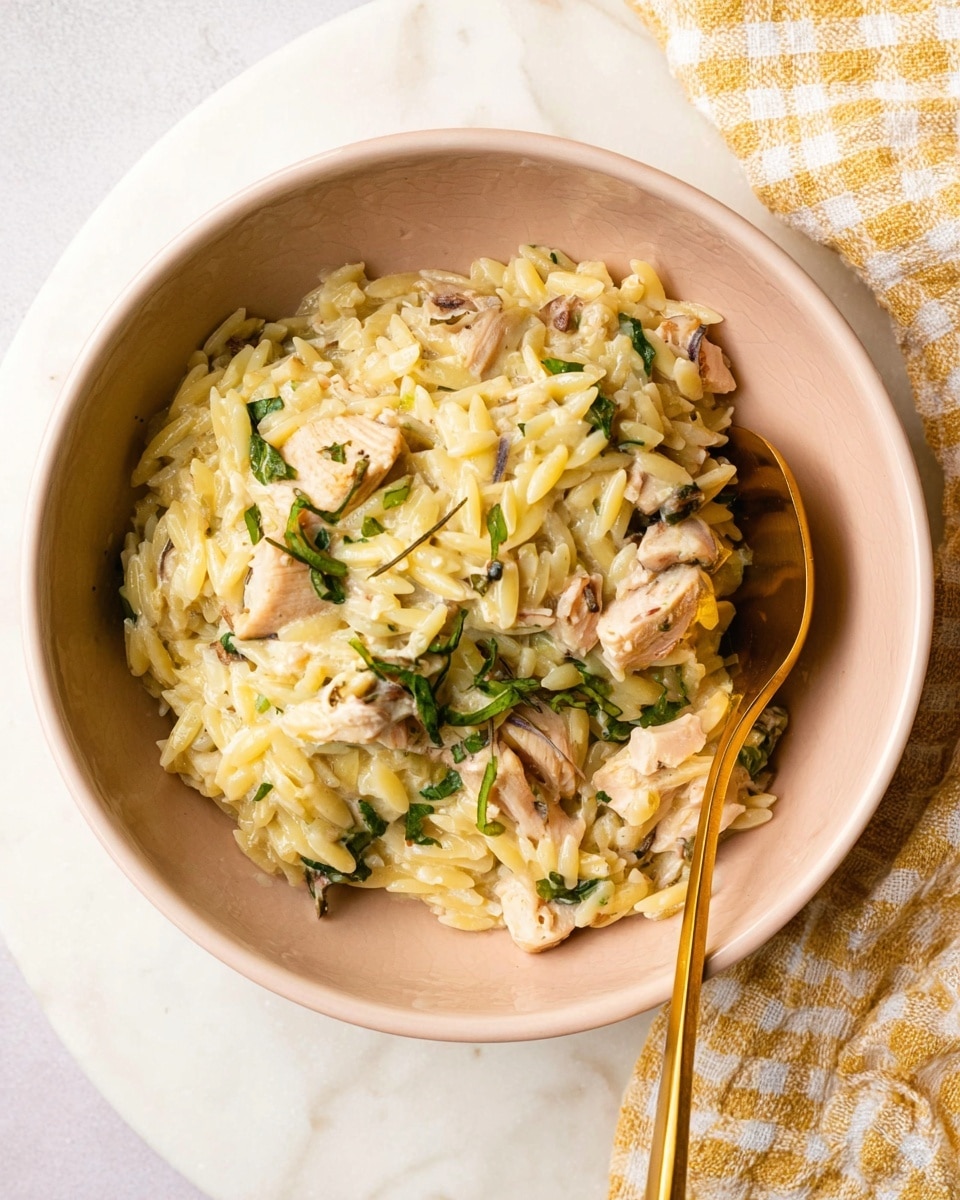 A close-up image of a creamy dish served in a light peach bowl, placed on a round white marbled surface. The dish has a base layer of yellowish creamy orzo pasta mixed with small bits of cooked chicken, which is light pinkish-beige in color and appears tender. There is a sprinkle of fresh green herbs on top, adding a pop of color. A gold spoon is partly inside the bowl on the right side, resting against the edge. A light yellow and white checkered cloth is placed next to the bowl in the upper right corner. photo taken with an iphone --ar 4:5 --v 7