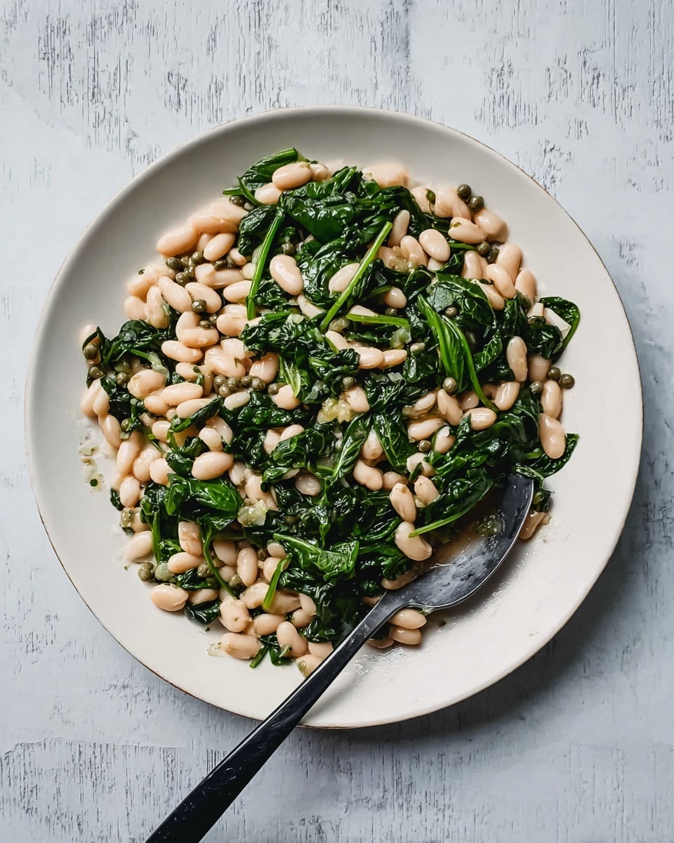 A large white plate holds a dish made of two main layers: a base layer of pale white beans scattered evenly across the plate, mixed with dark green, wilted spinach leaves that sit on top and throughout, giving a contrast of light and dark colors. Small green capers are mixed in with the beans and spinach, adding small dots of bright green. A silver spoon with a black handle rests on the edge of the plate, partly lying over the food. The whole plate sits on a white marbled surface, showing faint grey and beige patterns. photo taken with an iphone --ar 4:5 --v 7