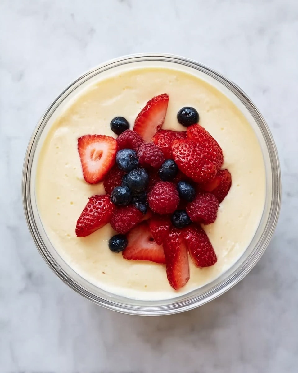 A clear glass bowl sits on a white marbled surface, filled mostly with a thick, pale yellow cream base that looks smooth and silky. On top, there is a pile of fresh berries: bright red strawberries sliced to show their juicy inside, small dark red raspberries, and round deep blue blueberries scattered together in the center. The colors of the berries contrast nicely with the creamy yellow beneath, and the glass bowl shows the cream’s thickness through its sides. photo taken with an iphone --ar 4:5 --v 7