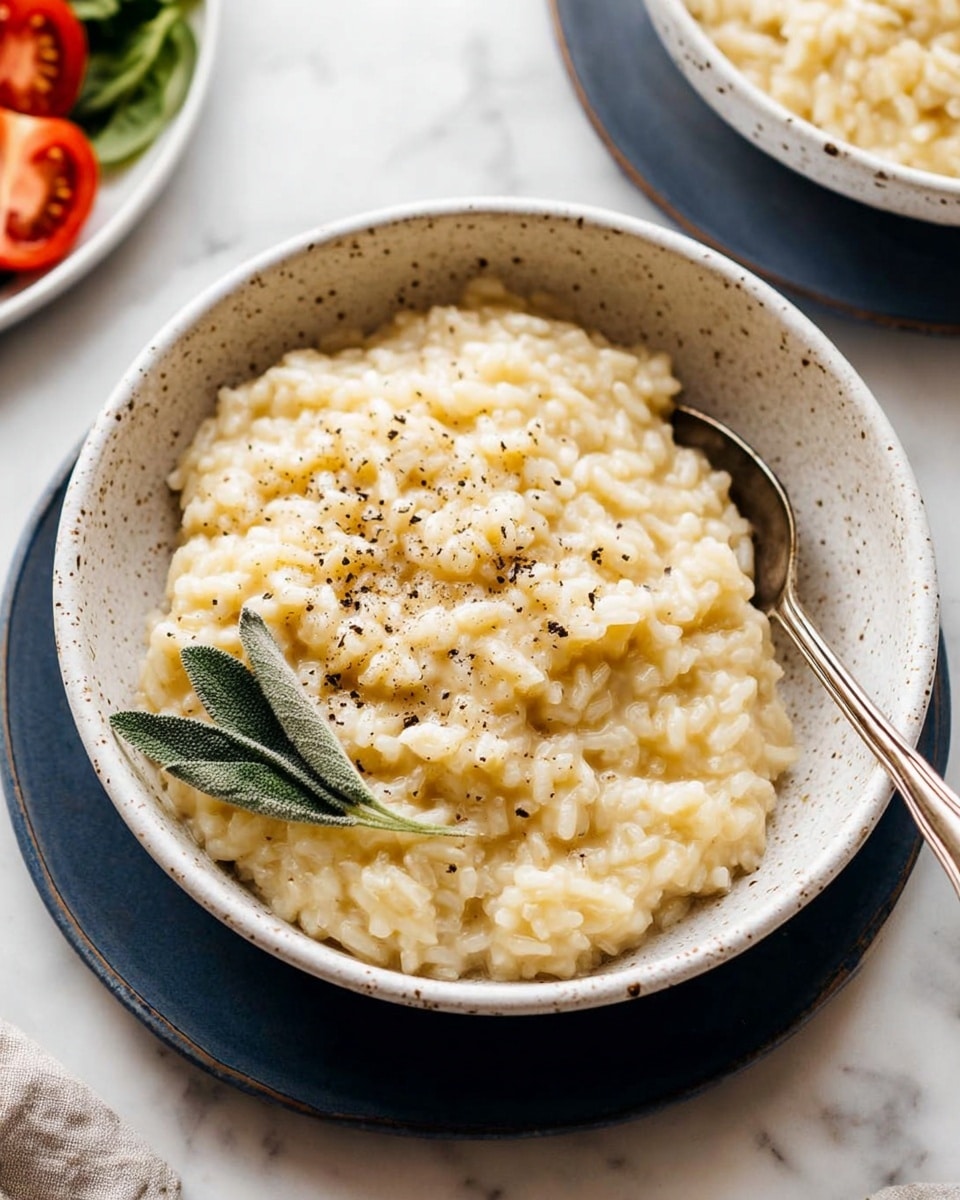 A close-up view of a white speckled bowl filled with creamy risotto, pale yellow and smooth with small grains of rice visible, topped with a sprinkle of black pepper and a small green sage leaf on the left side. The bowl sits on a dark blue plate, and a silver spoon rests inside the bowl on the right. The background features a white marbled surface with some green spinach and sliced tomato visible on the left edge. Photo taken with an iphone --ar 4:5 --v 7