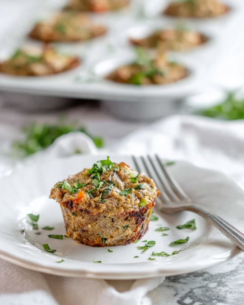 The image shows a small muffin-shaped dish with a rough and chunky texture, mainly light brown with some orange and green bits mixed inside, topped with small pieces of fresh green herbs scattered on and around it. The muffin sits on a white plate with a slightly wavy edge, placed on a white marbled surface. In the blurred background, there is a white muffin tray holding multiple similar muffins, also garnished with green herbs. A metallic fork rests on the right side of the plate. photo taken with an iphone --ar 4:5 --v 7