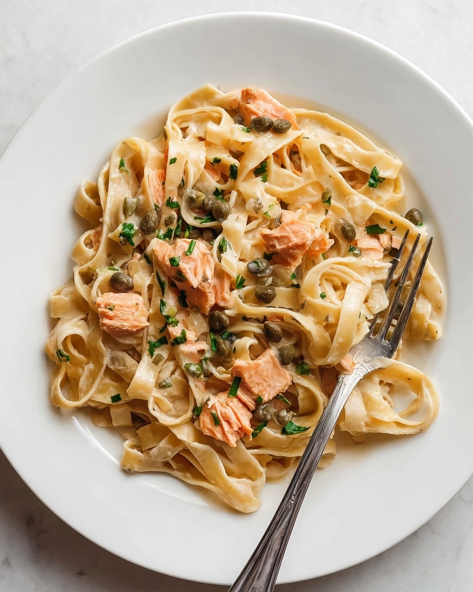 A white plate holds a creamy light beige pasta dish with flat, wide noodles arranged loosely in a circular pattern, coated evenly with sauce. Scattered on top are small pieces of soft pink salmon, small dark green capers, and finely chopped fresh green chives. A silver fork is tangled with some noodles near the bottom right of the plate. The plate sits on a white marbled surface. Photo taken with an iphone --ar 4:5 --v 7