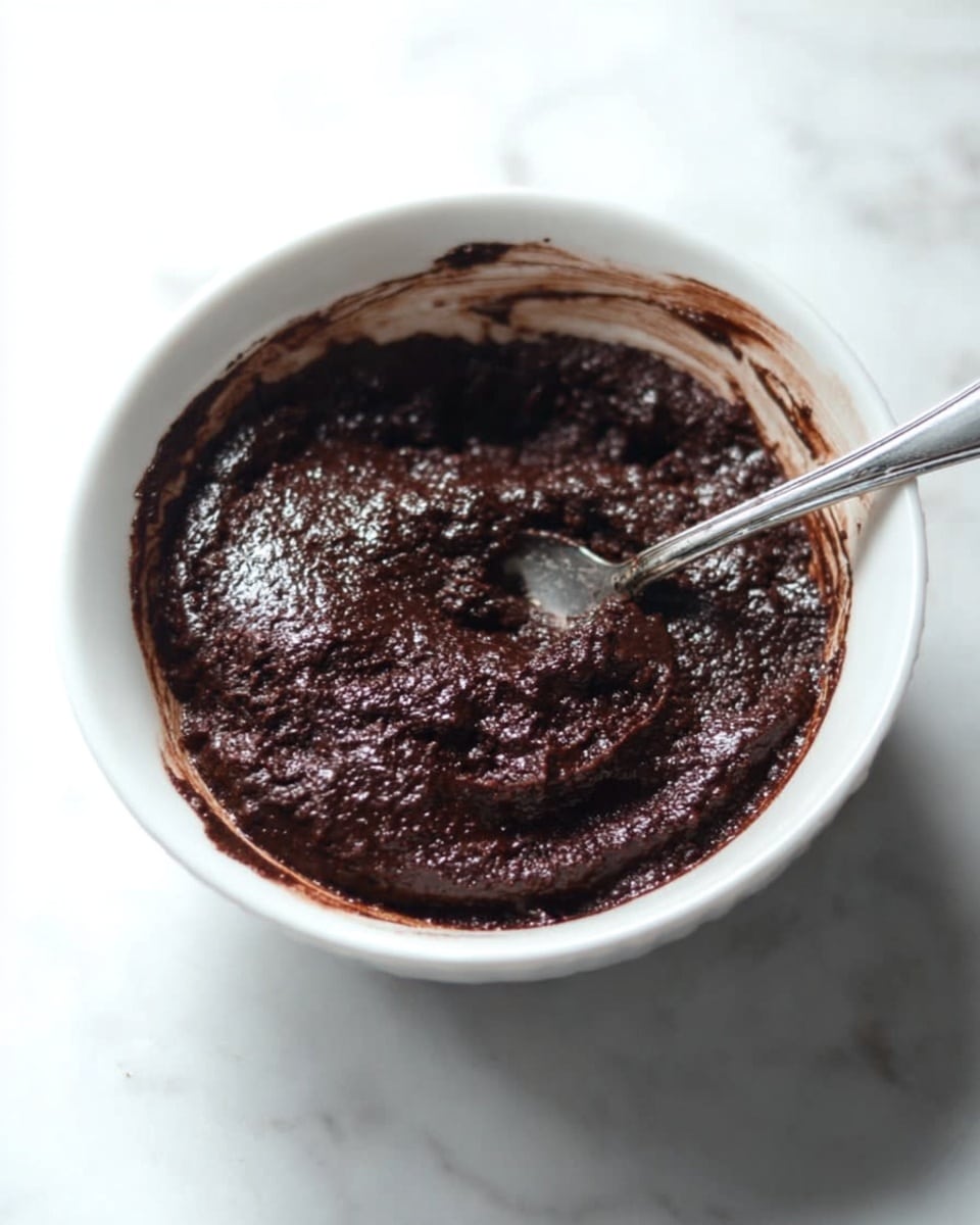 The image shows a white bowl filled with a thick, dark chocolate mixture that looks moist with a slightly bubbly, textured surface. A silver spoon rests inside the bowl, partly dipped into the rich, dense chocolate. The bowl is placed on a white marbled surface, enhancing the contrast with the dark brown dessert inside. The edges of the bowl have some chocolate smudges, showing it was stirred recently. photo taken with an iphone --ar 4:5 --v 7