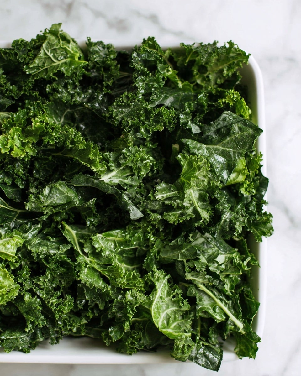 The image shows a close-up of a white rectangular dish filled with a thick layer of fresh kale leaves. The kale is a mix of dark green and lighter green shades with a curly texture, some leaves appearing glossy and wet while others are matte and crinkled. The leaves fill the dish completely, creating a dense, textured appearance that contrasts with the smooth, white dish edges. The background is a white marbled surface. Photo taken with an iphone --ar 4:5 --v 7