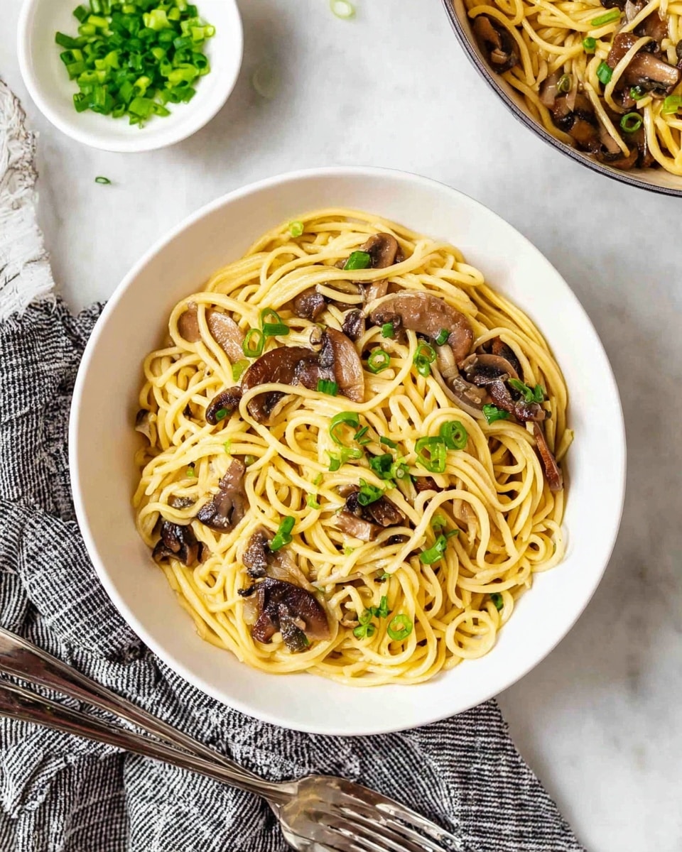 A white bowl filled with light yellow noodles mixed with dark brown sliced mushrooms, topped with small pieces of fresh bright green onions scattered evenly over the noodles. The bowl sits on a white marbled surface with a white dish holding more chopped green onions nearby. A checked cloth napkin in black and white lies below the bowl next to two shiny silver forks. Part of another white bowl with the same noodle and mushroom dish is partially visible in the top right corner. Photo taken with an iphone --ar 4:5 --v 7