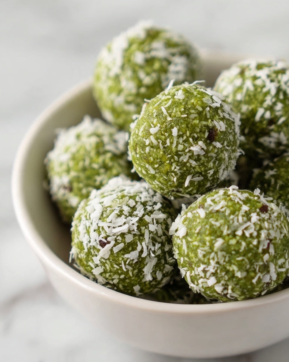 The image shows a close-up of a white bowl filled with round green balls that look like energy bites. Each ball is covered with small white coconut flakes and has a rough texture with some tiny dark seeds visible inside. The balls are stacked slightly uneven, with some overlapping each other, and the bowl sits on a white marbled surface. The focus is sharp on the balls in the center, showing their detailed texture. photo taken with an iphone --ar 4:5 --v 7