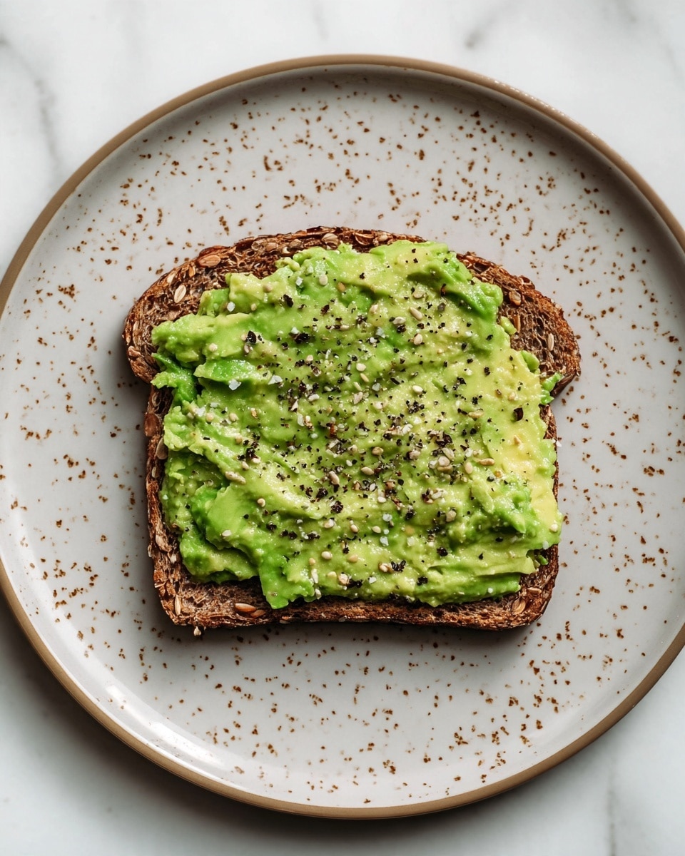 A single slice of brown multigrain toast with visible seeds and grains forms the base layer, topped thickly with a smooth, light green smashed avocado spread that has some small chunks for texture. The avocado layer is sprinkled evenly with coarse black pepper, adding contrast to the green color. The toast is placed on a white plate with tiny scattered brown specks, set against a white marbled background. photo taken with an iphone --ar 4:5 --v 7