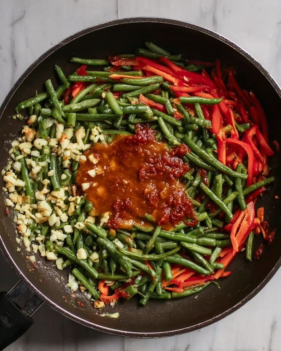A large dark pan contains three main layers: a base layer of small chopped garlic pieces scattered unevenly, a second layer of green beans cut into short pieces and red bell pepper strips spread around the pan, and a top layer of thick reddish-brown sauce in the center. The pan is placed on a white marbled surface. photo taken with an iphone --ar 4:5 --v 7