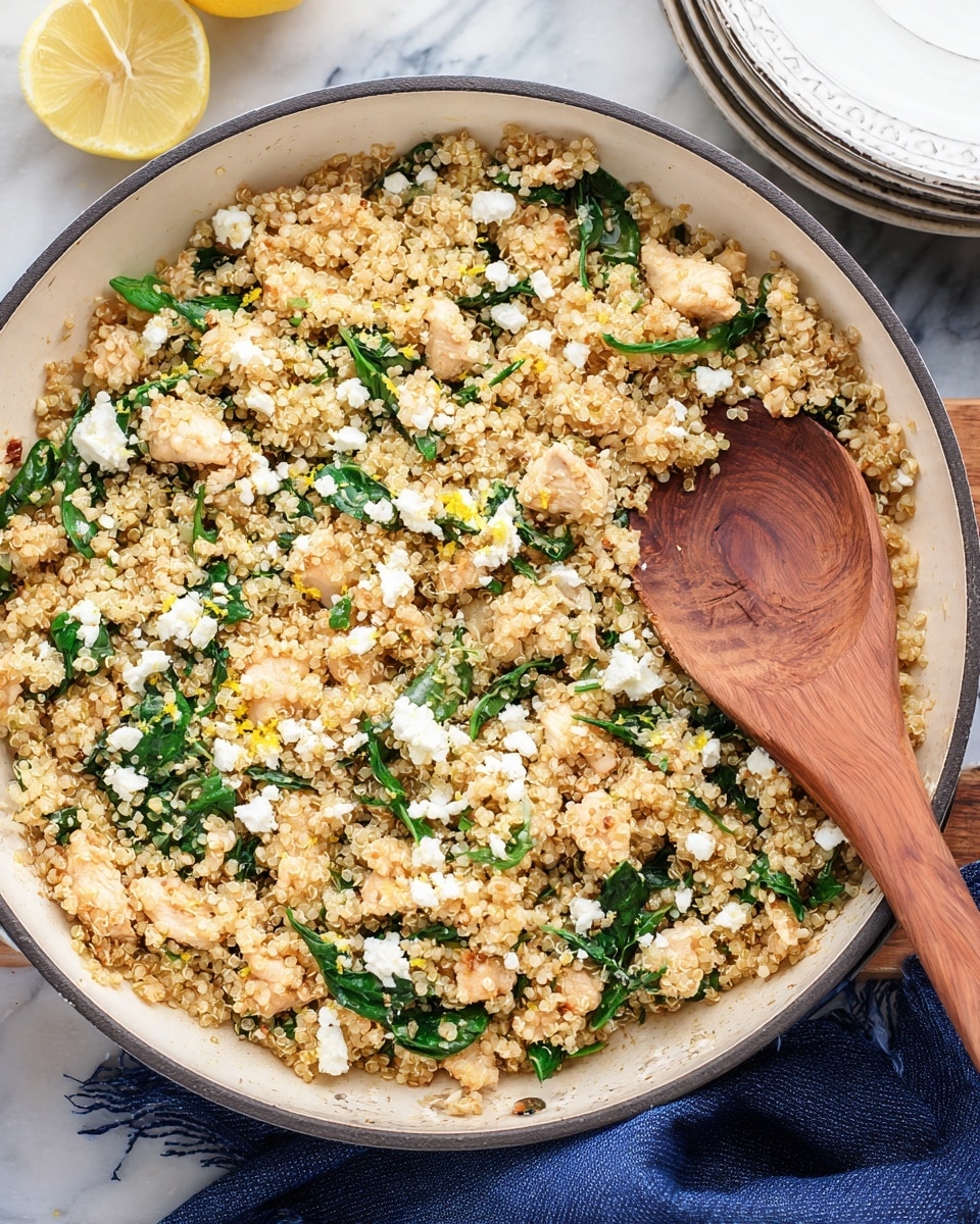 A close-up view of a white pan filled with a cooked quinoa dish that shows small, light golden quinoa grains mixed with small, light beige pieces of chicken, fresh dark green spinach leaves, white crumbled cheese dots, and small bits of yellow lemon zest spread evenly throughout. A wooden spoon lies on top of the quinoa mixture, showing its circular wood grain texture. The pan is set on a white marbled surface, with a folded dark blue cloth nearby, part of a stacked white plate, and a lemon wedge partially visible in the top corner. photo taken with an iphone --ar 4:5 --v 7