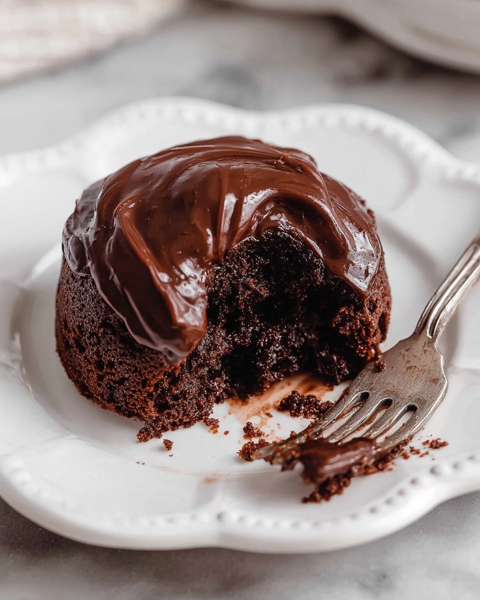 A single chocolate cake with one main thick layer sits on a white plate shaped with petal-like edges. The cake is covered with a glossy, rich chocolate frosting that swirls on top and drips a little down the sides. The middle of the cake has a small bite taken out, showing the dark, moist texture inside, with a bit of sauce pooled at the bottom. To the right of the cake is a silver fork with some crumbs stuck between its prongs. The plate is placed on a white marbled surface. photo taken with an iphone --ar 4:5 --v 7