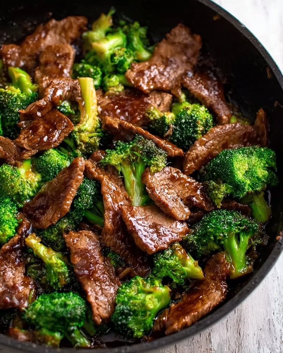 A close-up view of a dish in a black pan filled with thin slices of cooked beef that have a shiny, brown glaze, mixed with bright green steamed broccoli florets scattered evenly throughout. The beef pieces are layered on top and around the broccoli, with a slightly textured surface showing the cooking marks. The background beneath the pan is a white marbled texture. The rich colors of the beef and broccoli contrast nicely with the dark pan, showing a hearty and fresh stir-fry meal photo taken with an iphone --ar 4:5 --v 7