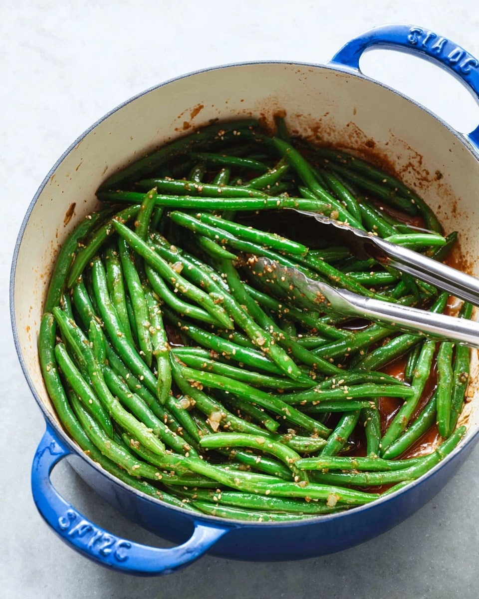 A blue pot with a white inside filled with bright green cooked green beans scattered evenly across the bottom, coated lightly in sauce with small bits of garlic and sesame seeds sprinkled throughout, and metal tongs resting on the right side inside the pot, all placed on a white marbled surface photo taken with an iphone --ar 4:5 --v 7