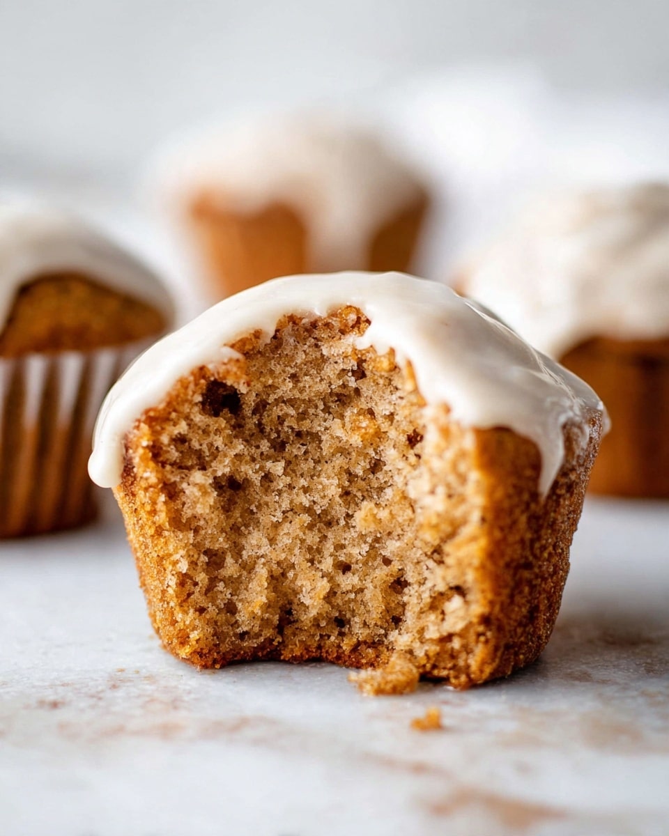 A close-up view of a single muffin with one thick layer, broken to show the soft and crumbly light brown inside texture. The muffin is topped with a smooth, white icing that drips slightly over its edge. In the background, there are two more whole muffins, out of focus, resting on a white marbled surface. Photo taken with an iphone --ar 4:5 --v 7