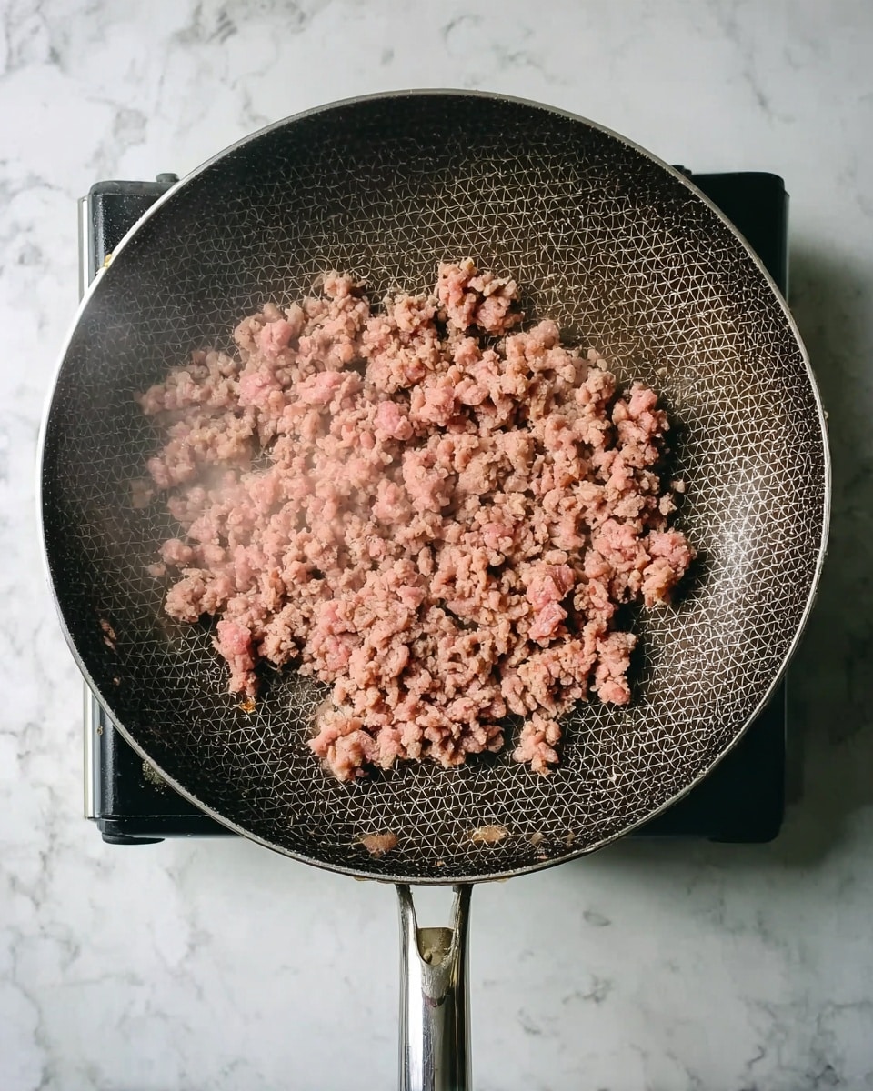 A round frying pan with a dark honeycomb pattern on the cooking surface contains a layer of ground meat cooking. The meat is unevenly spread, showing a mix of pink raw and light brown cooked parts, with some steam rising gently. The pan has a metallic handle and sits on a black electric stove top, placed on a white marbled surface. photo taken with an iphone --ar 4:5 --v 7