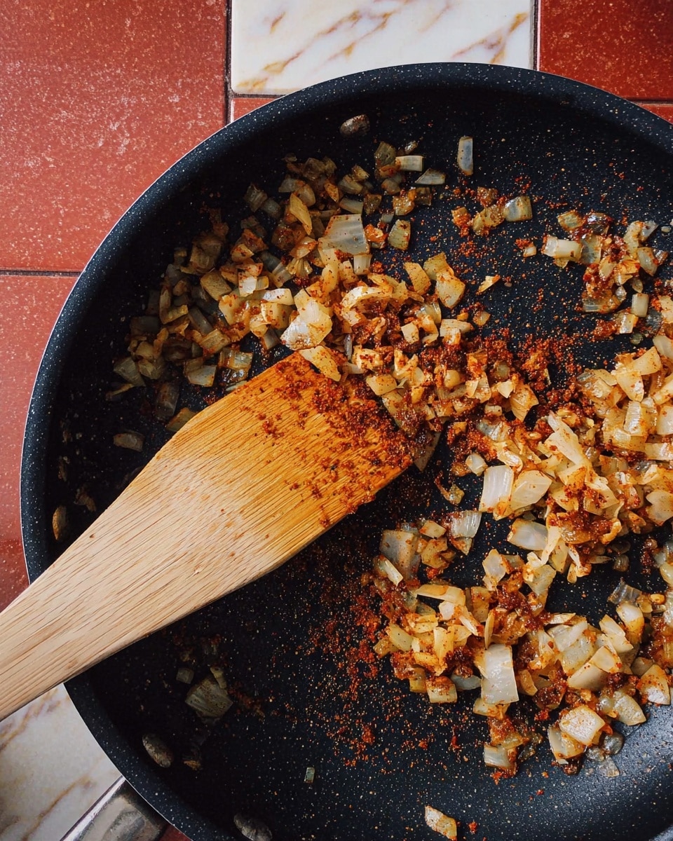 A black pan holds a mix of small pieces of cooked onion and garlic, each piece browned with a reddish spice coating, spread unevenly across the pan bottom. A light wooden spatula rests on top of the mixture, showing some spice stains on its edge. The background shows a white marbled surface partially covered by reddish tiles. photo taken with an iphone --ar 4:5 --v 7