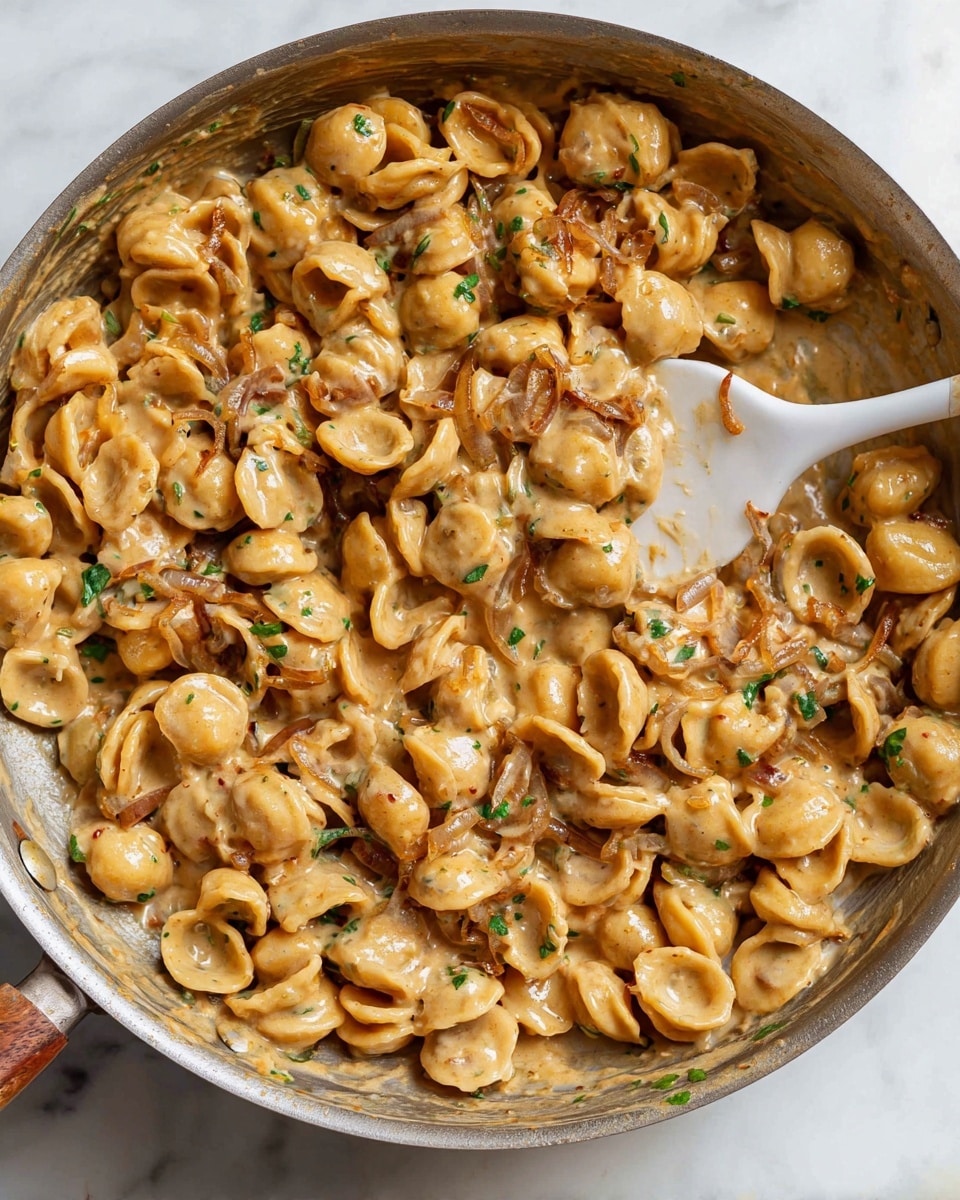 The dish shows a skillet filled with small round pasta pieces mixed with a creamy light brown sauce, making the pasta look smooth and coated. There are visible thin slices of light brown onion and flecks of green herbs sprinkled evenly throughout the dish. The sauce looks rich and slightly thick, clinging to each pasta piece and the onions. A white spatula with a wooden handle is inside the skillet, lifting some of the pasta, with a few pieces sticking to it. The skillet sits on a white marbled surface, highlighting the warm colors of the pasta and sauce. photo taken with an iphone --ar 4:5 --v 7