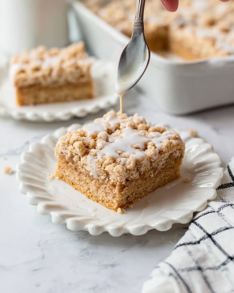 A single large square crumb bar sits on a white scalloped plate, showing two visible layers: a light beige dense base with a thick crumbly golden brown topping made of small uneven clusters. A spoon held by a woman's hand drizzles a thin white glaze over the top layer, making some moist spots visible on the crumb surface. Behind, a white rectangular pan contains more crumb bars, and a second crumb bar is on another white scalloped plate slightly out of focus. Everything rests on a white marbled surface, with a white and black checked cloth partially visible at the bottom right. Photo taken with an iphone --ar 4:5 --v 7