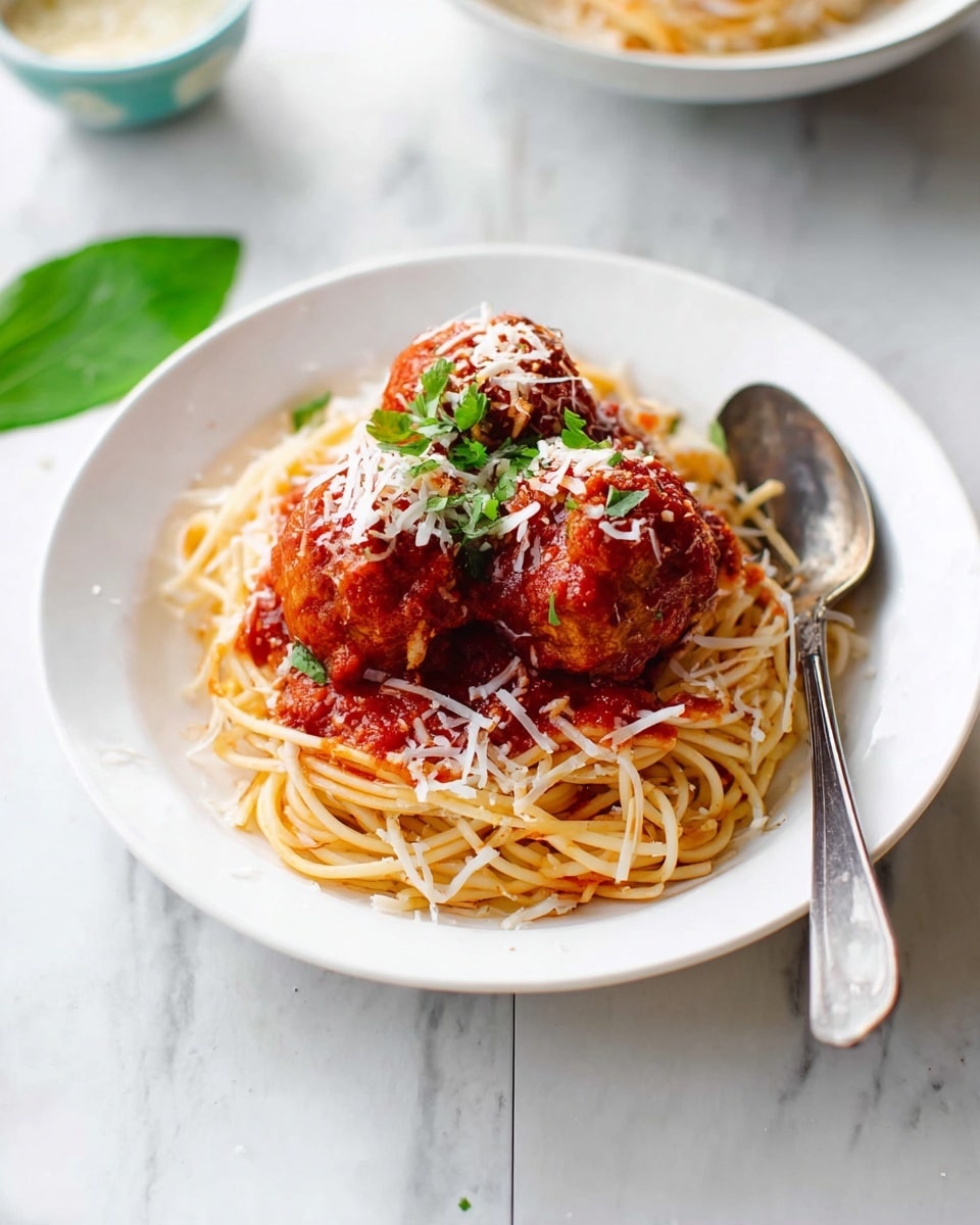 A white plate holds a neat nest of spaghetti with a light golden color, topped with three large reddish-brown meatballs covered in thick, bright red tomato sauce. The sauce spreads slightly over the edges of the pasta. White shredded cheese is sprinkled over the meatballs and sauce, adding a soft texture. A small green herb garnish sits on top of the meatballs, providing a fresh contrast. A fork and spoon rest on the right side of the plate. The background is a white marbled surface with a faint green leaf and a blurry small bowl behind the plate. Photo taken with an iphone --ar 4:5 --v 7