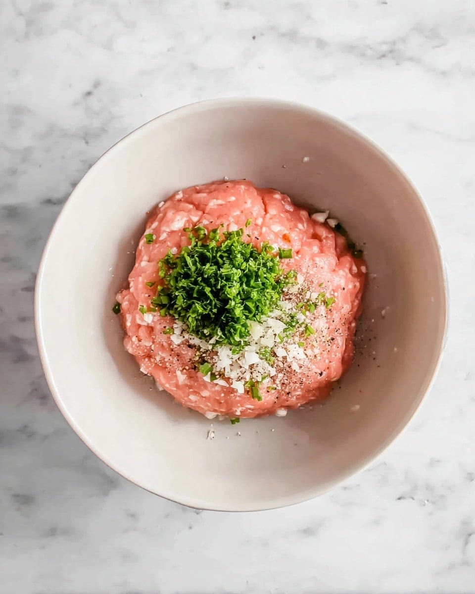 A white bowl with one large round layer of smooth light pink ground meat in the center. On top of the meat is a small pile of chopped green herbs, tiny pieces of white garlic, a sprinkle of white salt, and some black pepper. The bowl sits on a white marbled surface. photo taken with an iphone --ar 4:5 --v 7