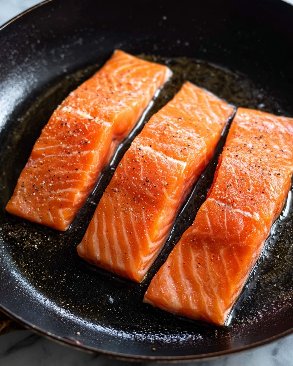 In this image, there are three pieces of fresh, bright orange salmon fillets placed side by side in a dark black pan. Each piece is thick and slightly shiny with natural salmon lines and a sprinkle of black pepper and salt on top. The surface of the salmon looks smooth and moist, showing clear texture from the fish's flesh. The pan has a slightly oily sheen, reflecting light softly around the salmon. The background is a white marbled texture. photo taken with an iphone --ar 4:5 --v 7