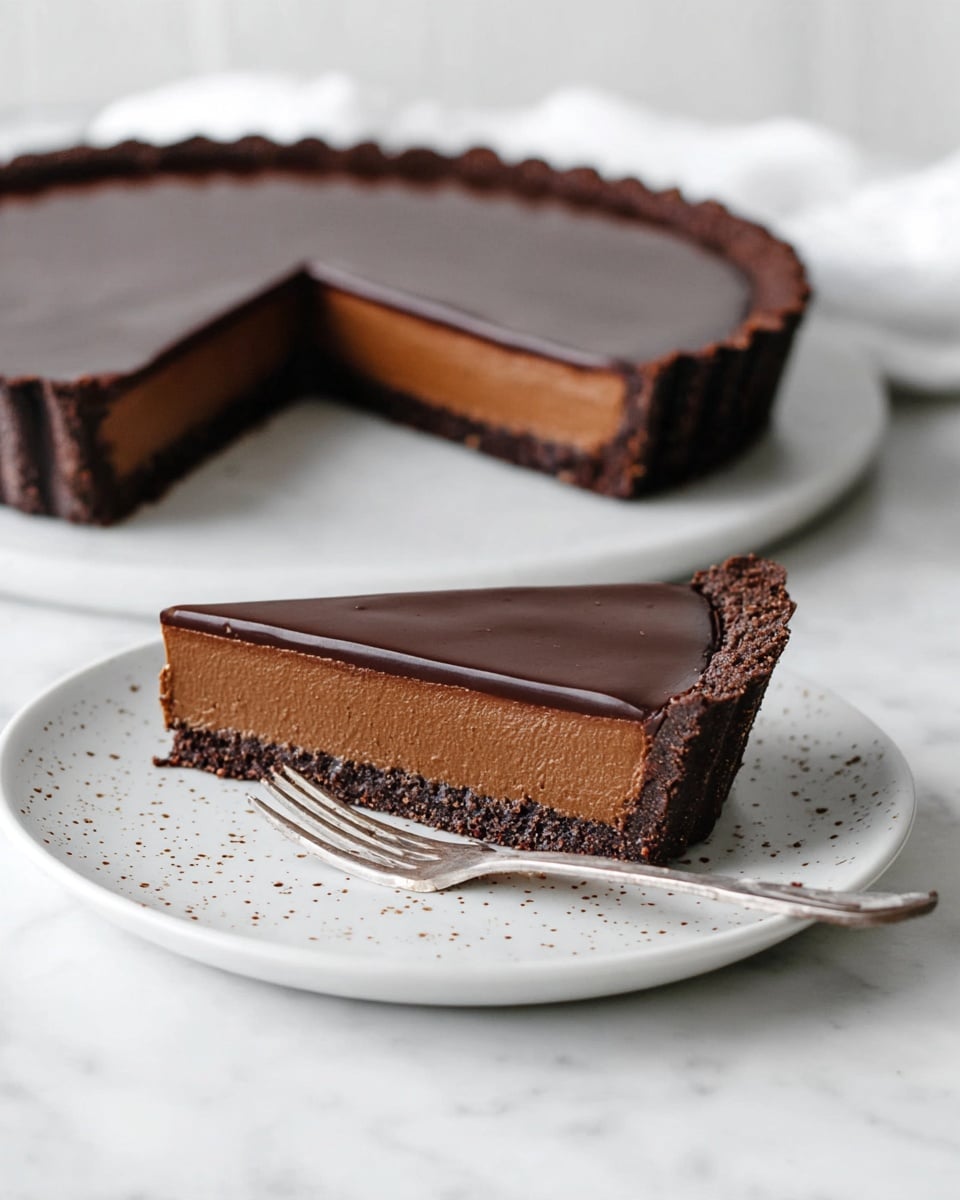 A smooth chocolate tart with three visible layers sits on a white marbled surface; the bottom layer is a firm, dark chocolate crust with a crumbly texture that holds the tart together, the middle layer is a thick, creamy milk chocolate filling with a slightly lighter brown color and soft texture, and the top layer is a glossy, dark chocolate ganache that is smooth and shiny. A slice is cut out and placed on a white plate with tiny brown specks, showing the same three layers clearly. A silver fork rests next to the slice on the plate, adding a simple touch. The background remains clean and minimalist with a white marbled texture. Photo taken with an iphone --ar 4:5 --v 7