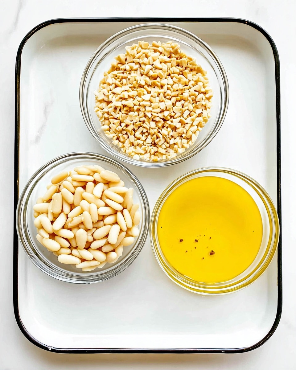 Three clear glass bowls sit on a white tray with a black rim, placed on a white marbled surface. The top left bowl is filled with small, light beige chopped nuts, showing a rough, uneven texture. To the right, another bowl contains smooth, oval-shaped pine nuts, creamy white with a slight shine. Below these, the third bowl holds a bright yellow liquid, smooth with a few small bubbles, likely melted butter or oil. The arrangement is simple and clean with a bright, natural light. photo taken with an iphone --ar 4:5 --v 7