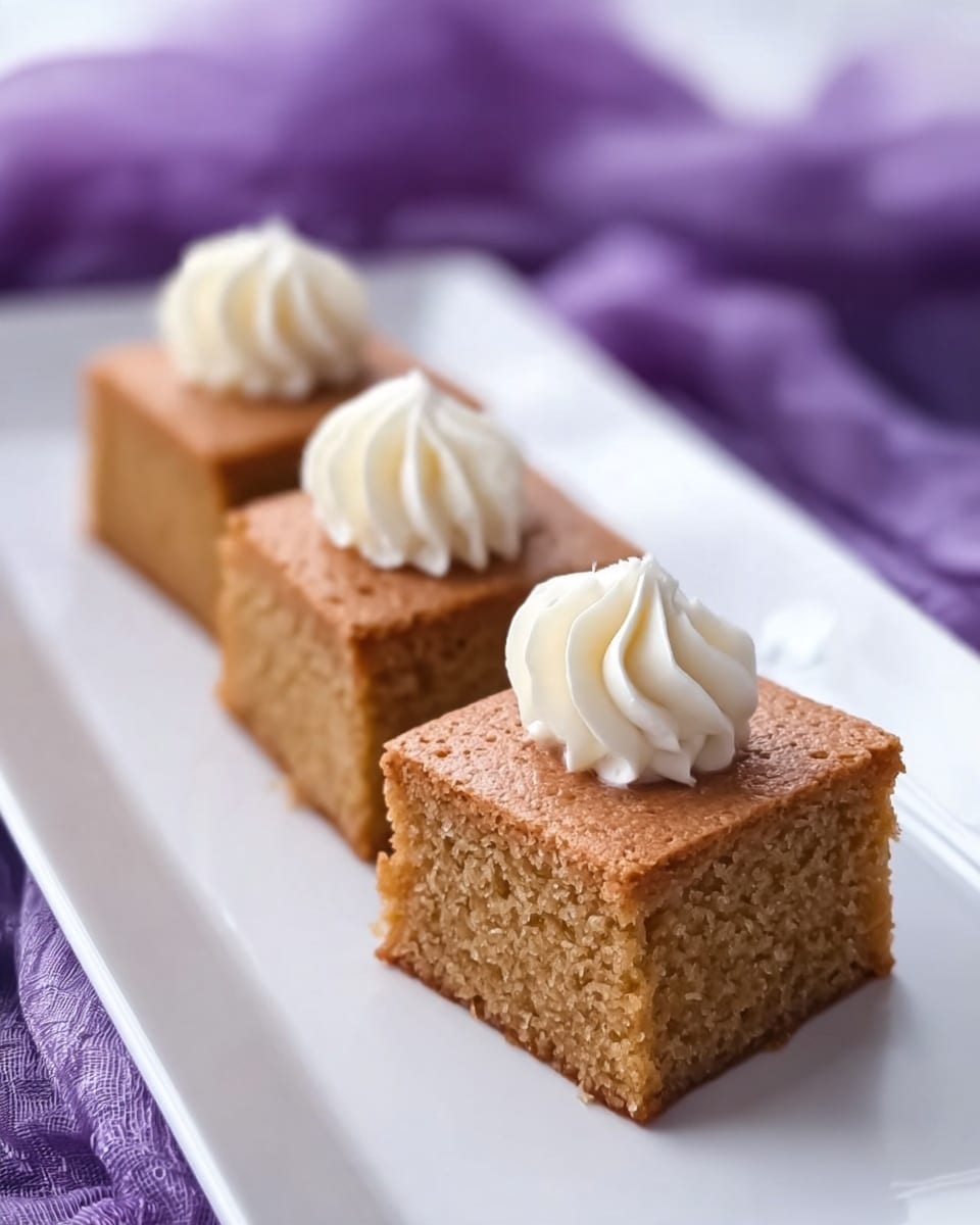 The image shows three square pieces of light brown cake arranged in a row on a long white plate with a white marbled surface underneath. Each cake piece has three small, round dollops of white cream frosting on top, placed close together near one corner, with the frosting having a ridged, piped texture. The background features blurred folds of a purple cloth around the plate. Photo taken with an iphone --ar 4:5 --v 7