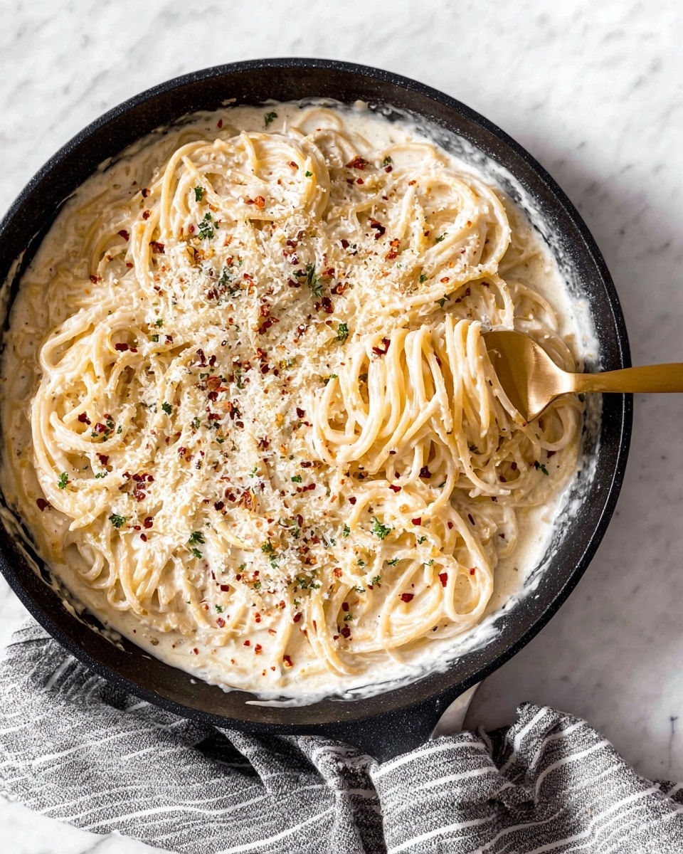 A black skillet filled with creamy spaghetti covered in a thick white sauce. The spaghetti strands are mixed in the sauce, with some lifted by a gold fork on the right side. The top layer is sprinkled with grated white cheese, black pepper, and small red chili flakes. The skillet is placed on a white marbled surface with a gray and white striped cloth wrapped around its handle. Photo taken with an iphone --ar 4:5 --v 7