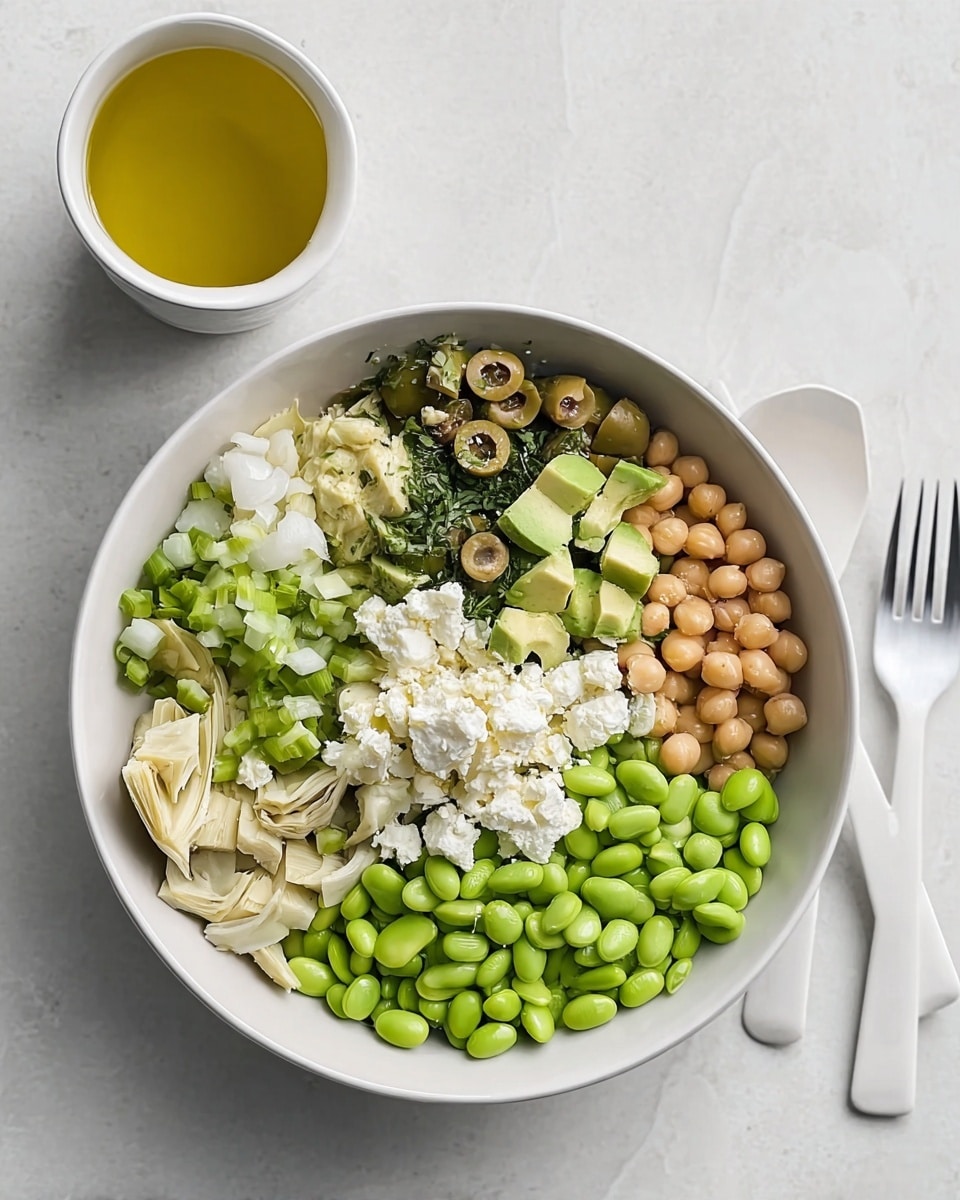 Inside a white bowl on a white marbled surface, there are eight visible layers arranged side by side by side. Starting from the bottom left going clockwise: light beige artichoke hearts with a soft texture, bright green diced avocado with a smooth texture, diced green celery, chunky white cheese crumbles, sliced green olives, a cluster of dark green pumpkin seeds, small round tan chickpeas, and a section of chopped fresh green herbs with additional white cheese crumbles on top. At the center bottom is a pile of shiny bright green edamame beans with a smooth surface. To the right of the bowl, two white utensils are placed on the white marbled surface, and above them is a white cup with yellow olive oil inside. photo taken with an iphone --ar 4:5 --v 7