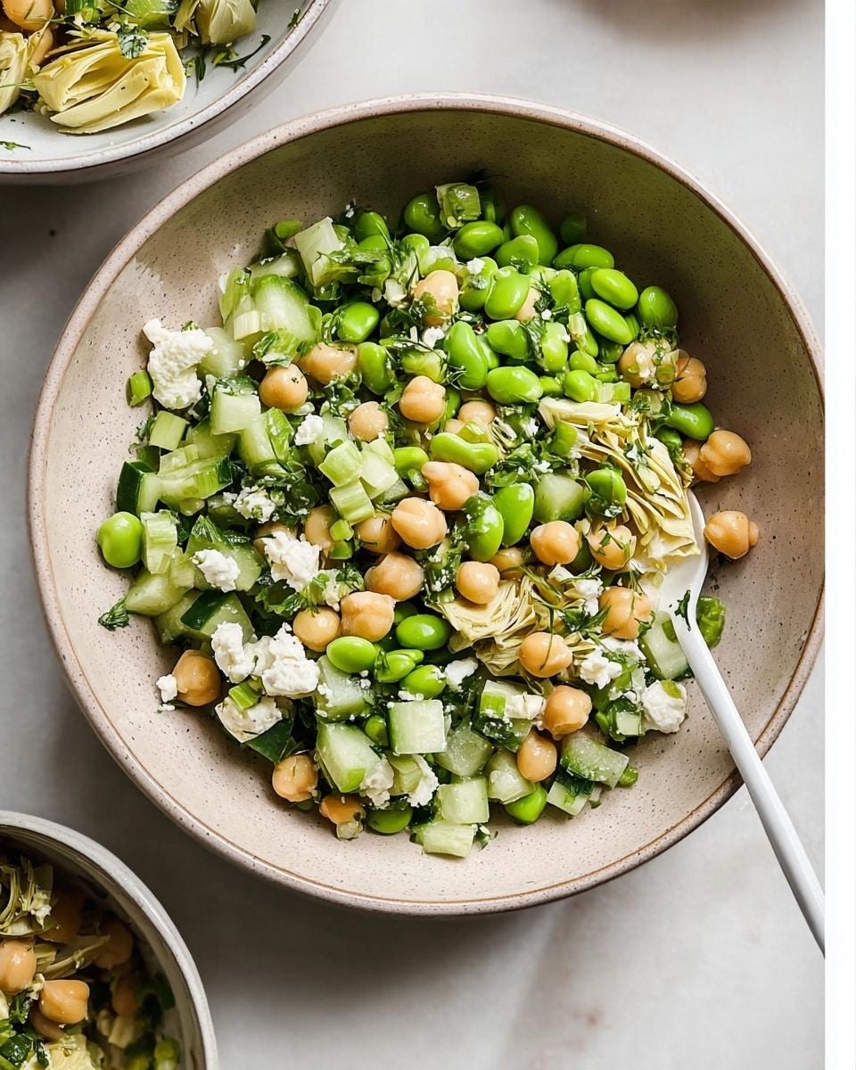 A beige bowl filled with a layered salad starting with a base of light green diced cucumber and celery pieces, topped with soft beige chickpeas and bright green edamame beans. Scattered on top are small chunks of creamy white cheese and pale artichoke hearts mixed with fresh green herbs, creating a fresh and colorful look. A white spoon rests inside the bowl on the right side. The bowl sits on a white marbled surface with parts of other bowls partially showing around the edges. photo taken with an iphone --ar 4:5 --v 7