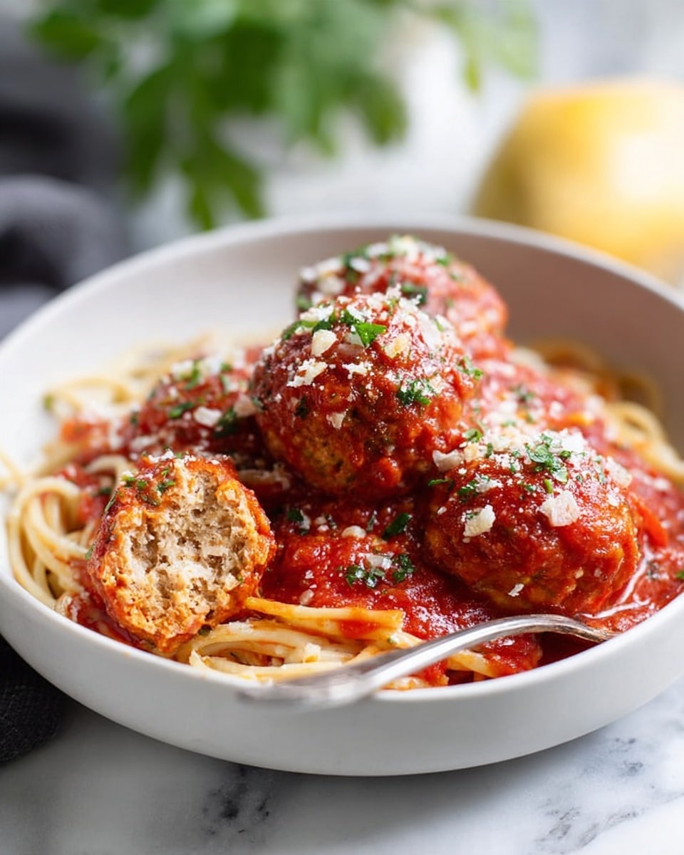 A white bowl holds a dish of spaghetti topped with three large meatballs covered in bright red tomato sauce. The spaghetti looks soft and is coated lightly with sauce at the base. One meatball is cut open, showing a light, textured inside with visible small bits. The meatballs are sprinkled with small green herb pieces and grated cheese on top. A silver fork rests inside the bowl near the cut meatball. The bowl sits on a white marbled surface with a blurred green plant and yellow item in the background. photo taken with an iphone --ar 4:5 --v 7