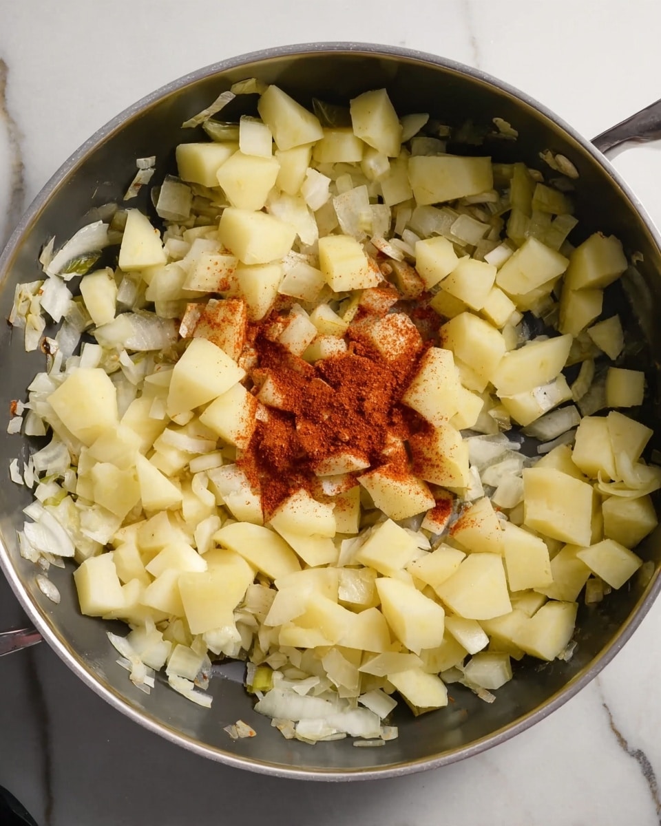 A large silver pan is filled with a mixture of chopped white onions and pale yellow potato pieces. The onions form a thin, translucent layer scattered across the bottom and sides of the pan, while the potato chunks are placed on top, mostly in the center. In the middle, a small pile of reddish-brown spice powder lies over the potatoes, adding a bright contrast to the muted colors. The pan sits on a white marbled surface, and no other objects are visible. Photo taken with an iphone --ar 4:5 --v 7