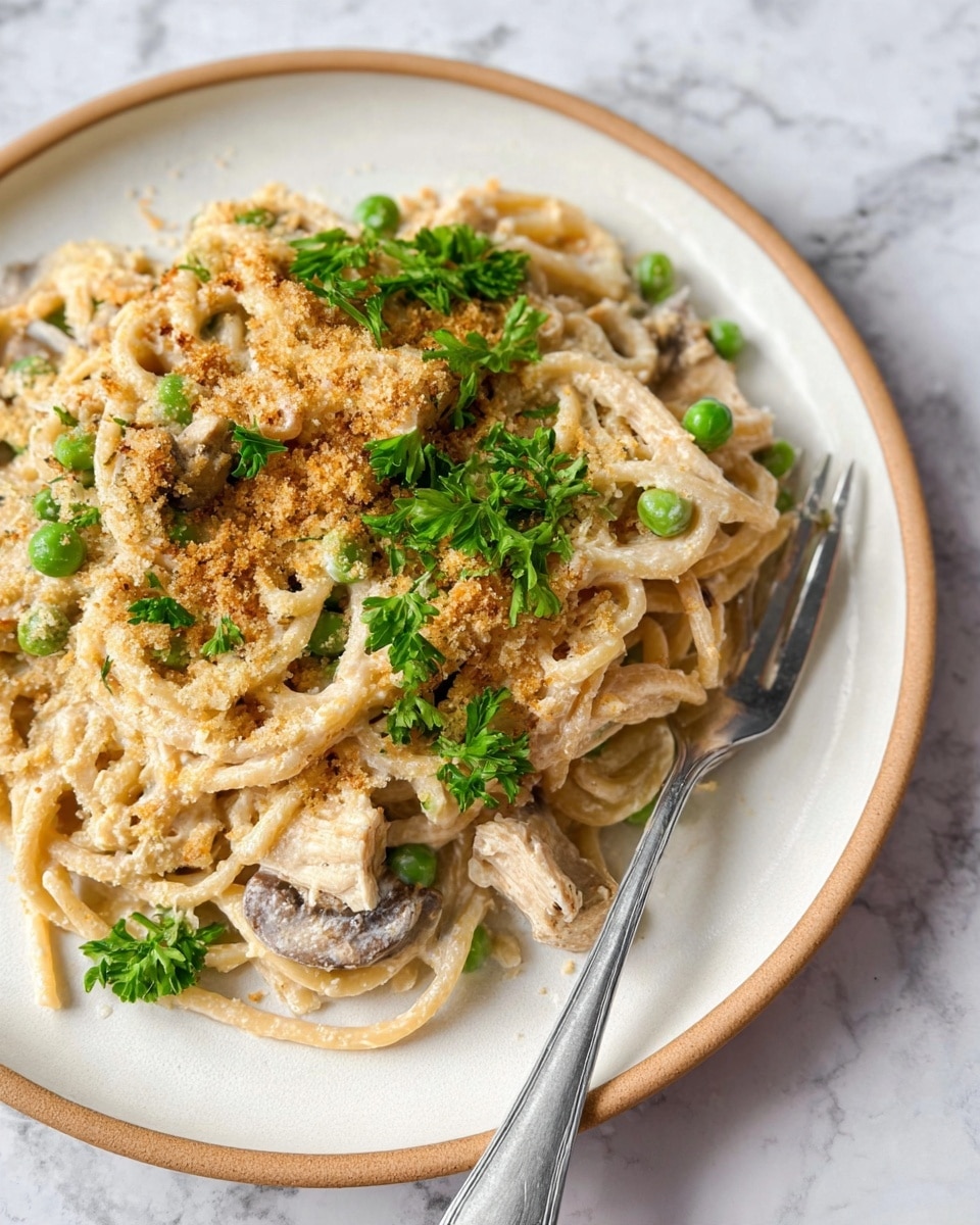 A close-up view of a creamy pasta dish served on a white plate with a light brown rim, placed on a white marbled surface. The dish features three main layers: at the bottom, a tangle of beige, creamy noodles mixed with green peas and small bits of chicken; the middle layer shows slices of light brown mushrooms and a sprinkle of golden brown breadcrumbs; on top, fresh bright green parsley leaves scattered over the pasta, adding a pop of color. A silver fork rests on the right side of the plate, slightly embedded in the pasta. Photo taken with an iphone --ar 4:5 --v 7