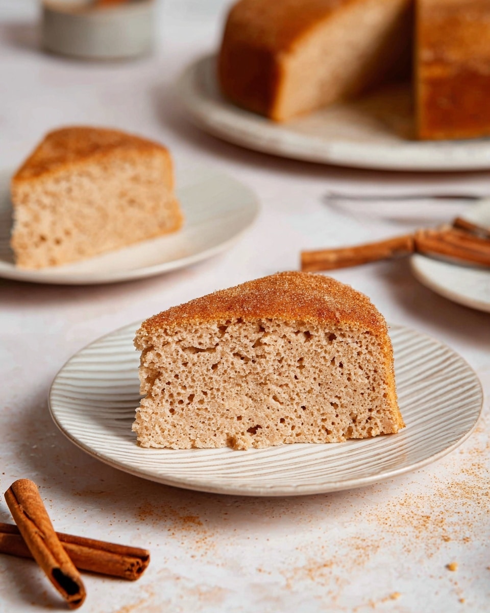 A slice of spongy, light brown cake with a rough texture and tiny holes is placed on a white plate with a subtle striped pattern. The cake slice has one single thick layer with a slightly darker brown, cinnamon-sugar sprinkled top. In the background, there are more slices and a larger cake on blurred white plates, all sitting on a white marbled surface. Two cinnamon sticks lie beside the foreground plate. The scene is softly lit, giving a cozy and warm feel. photo taken with an iphone --ar 4:5 --v 7