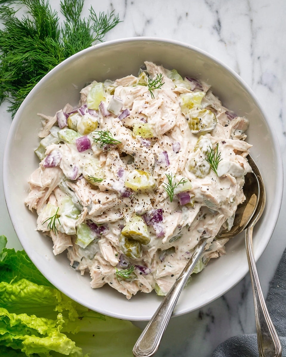 A white bowl filled with a creamy salad made of shredded pale pink chicken layers, small diced purple onions, and green pickle chunks, all mixed in a smooth white mayonnaise-like dressing. The salad is sprinkled with small green dill leaves and a light dusting of black pepper on top. Two silver spoons rest inside the bowl, slightly submerged in the salad. Around the bowl, there are fresh green lettuce leaves and sprigs of dill on a white marbled surface. Photo taken with an iphone --ar 4:5 --v 7