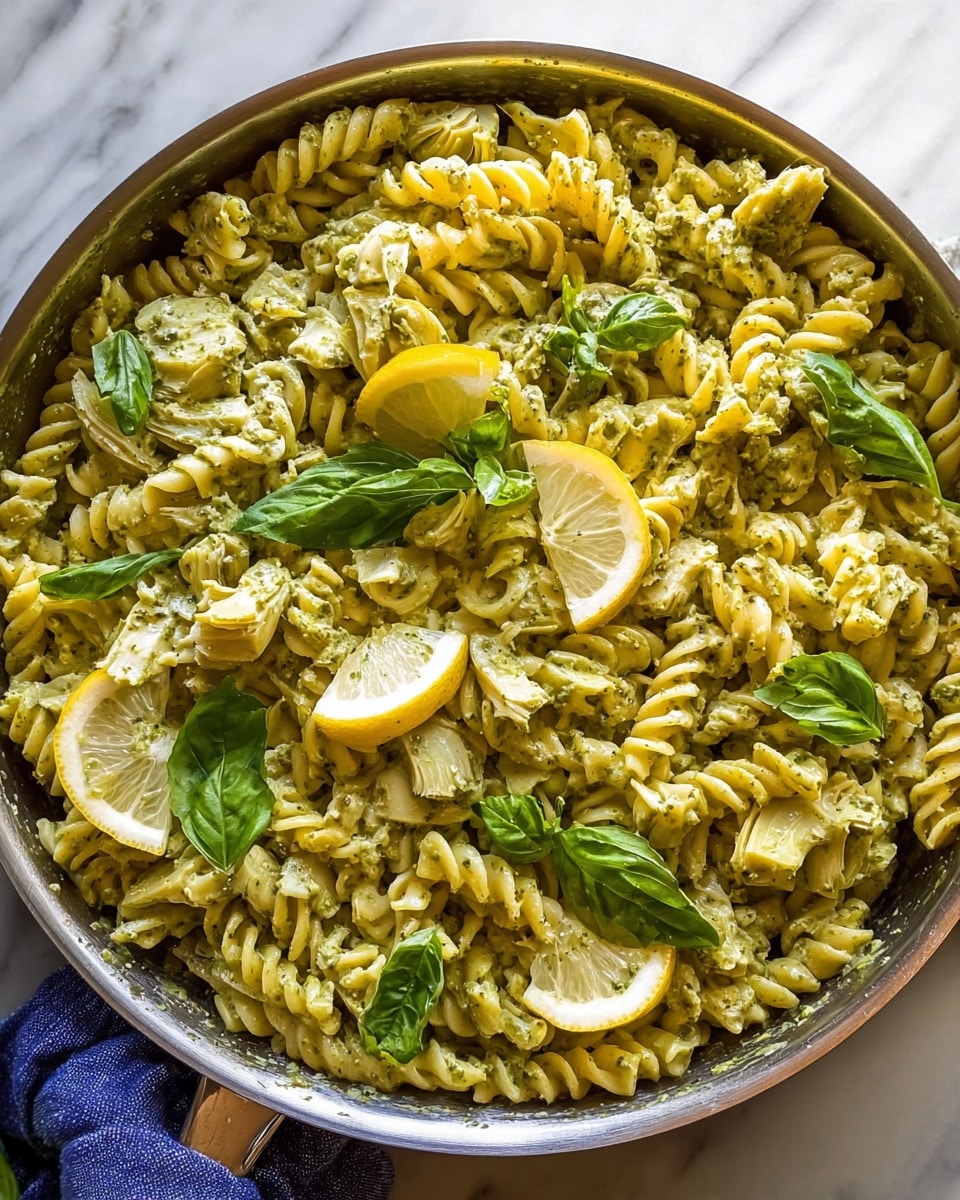 A large round pan holds a pasta dish with about three layers: the bottom layer is yellow twisted rotini pasta, covered in a light green pesto sauce giving a creamy texture; the second layer has pieces of light green artichoke hearts mixed evenly with the pasta; on top, scattered fresh green basil leaves add color and freshness; three yellow lemon wedges with white inner flesh rest near the center, slightly raised above the pasta. The pan sits on a white marbled surface with a small part of a blue cloth visible at the bottom left. photo taken with an iphone --ar 4:5 --v 7