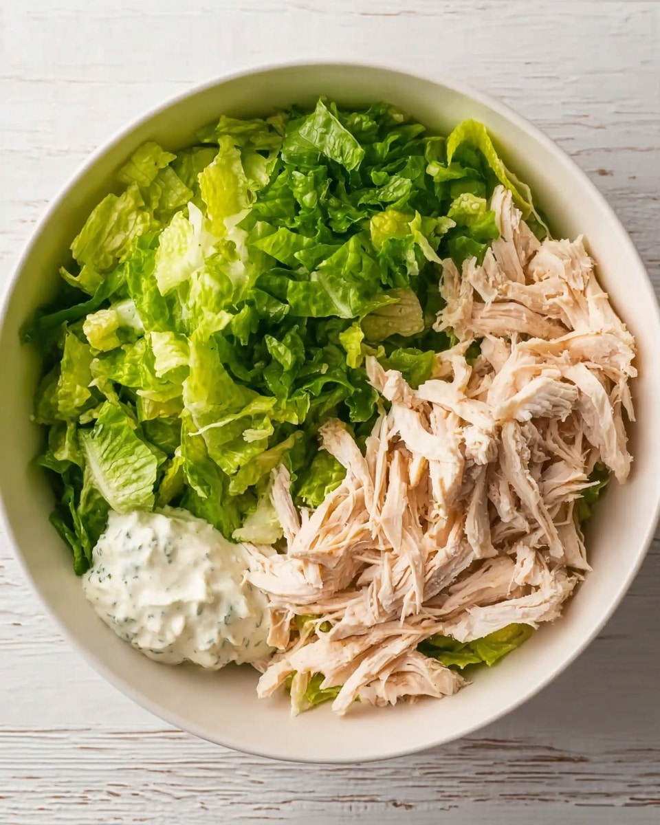 A white bowl contains three visible layers: fresh green chopped lettuce on the left side, creamy white dressing in a small dollop near the lettuce, and light beige shredded chicken piled on the right side. The bowl is placed on a white marbled surface with light wood texture showing underneath. The image has a simple and clean look, focusing on the fresh ingredients. Photo taken with an iphone --ar 4:5 --v 7