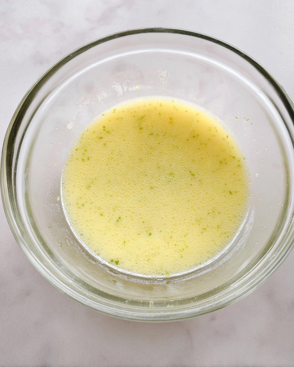 A clear glass bowl holds a single layer of light yellow liquid with tiny green specks mixed throughout. The liquid has a smooth texture with small bubbles on its surface, and the bowl is placed on a white marbled surface. The inside edges of the glass bowl show faint smudges of the liquid, giving a sense of recent stirring or mixing. Photo taken with an iphone --ar 4:5 --v 7